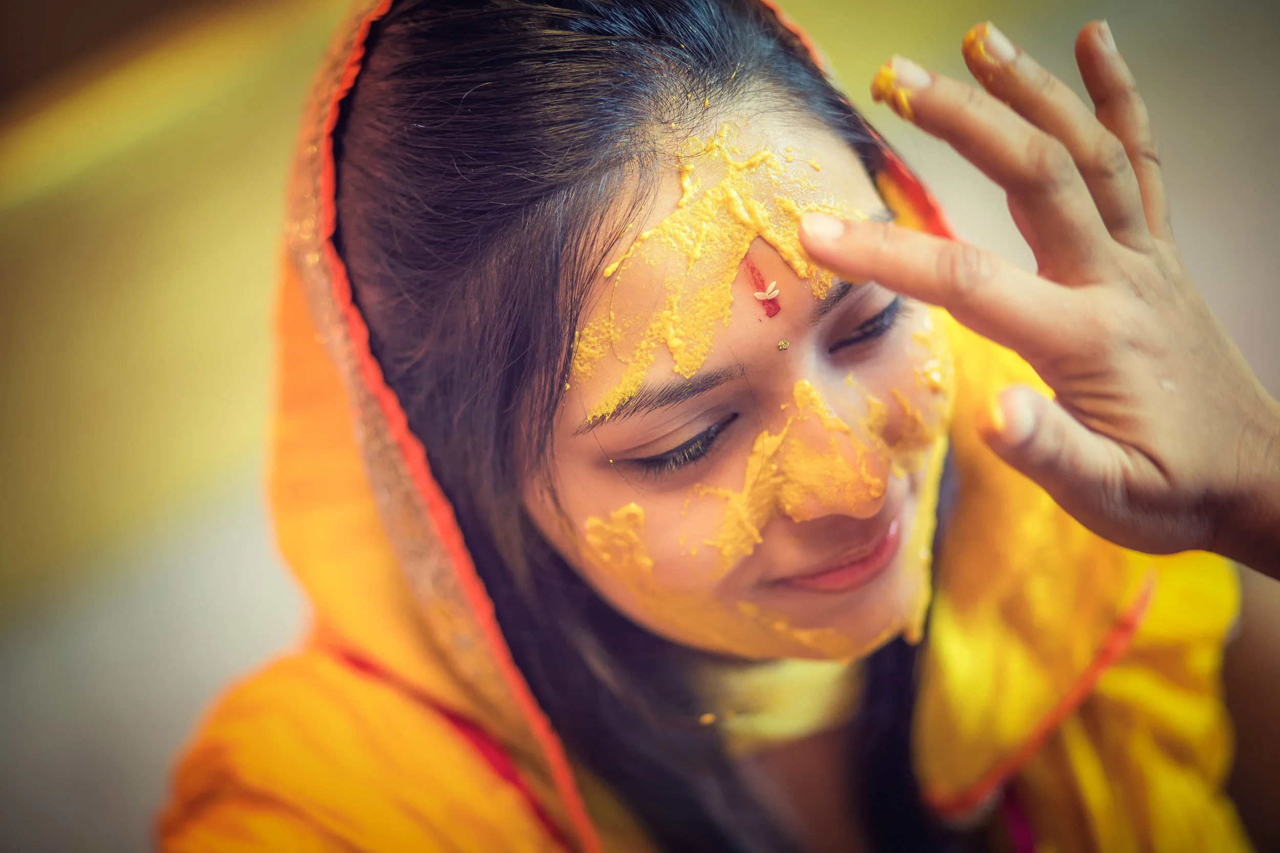 A woman with yellow powder on her face, wearing a yellow hoodie with orange trim, in a close-up shot with her eyes closed and her hand near her face.