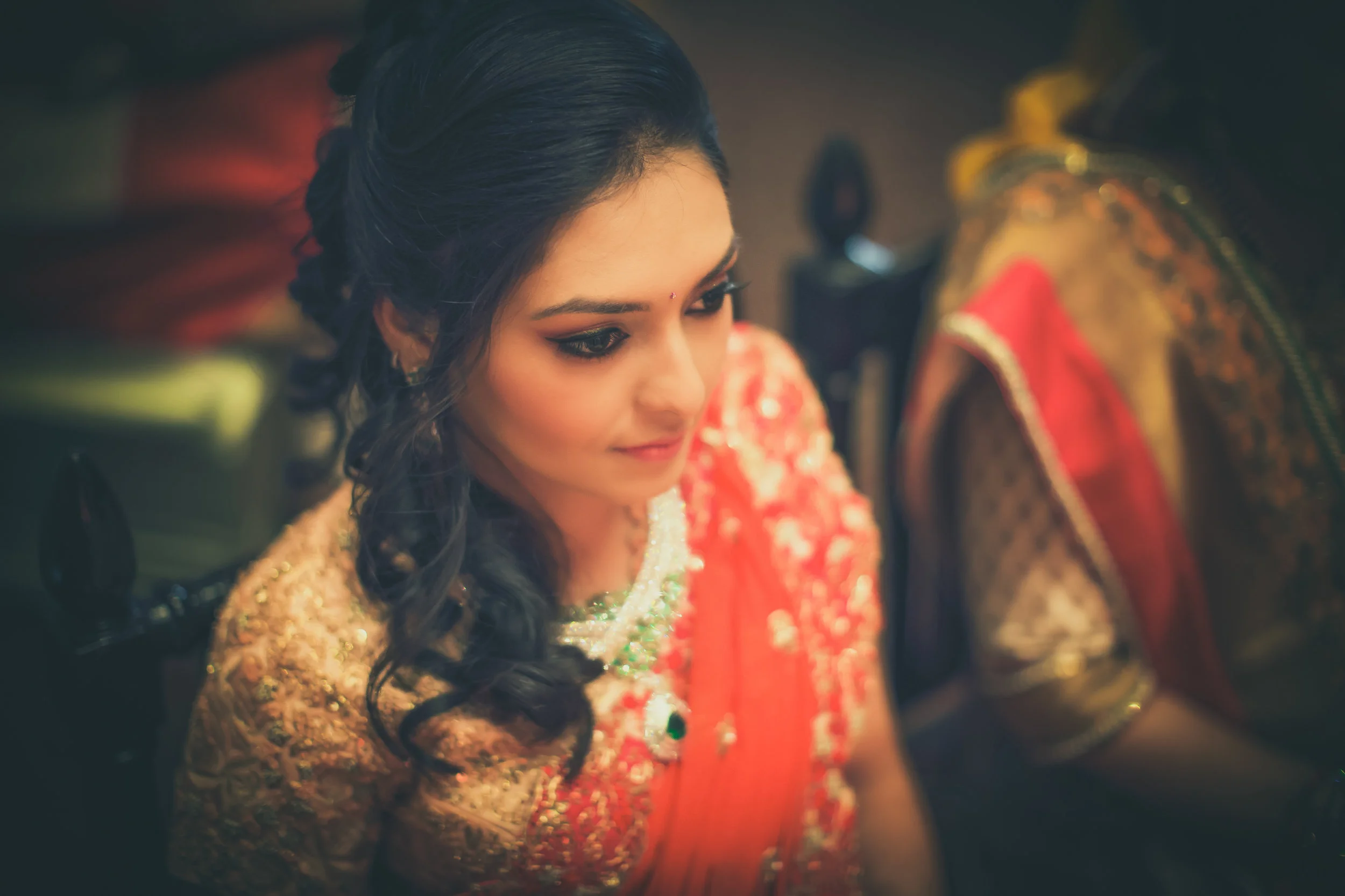 A woman dressed in traditional Indian attire, with dark wavy hair, is looking slightly downward. She is wearing an ornate, red and gold embroidery outfit, and jewelry. The background shows a traditional setting with colorful fabric and decorative ite