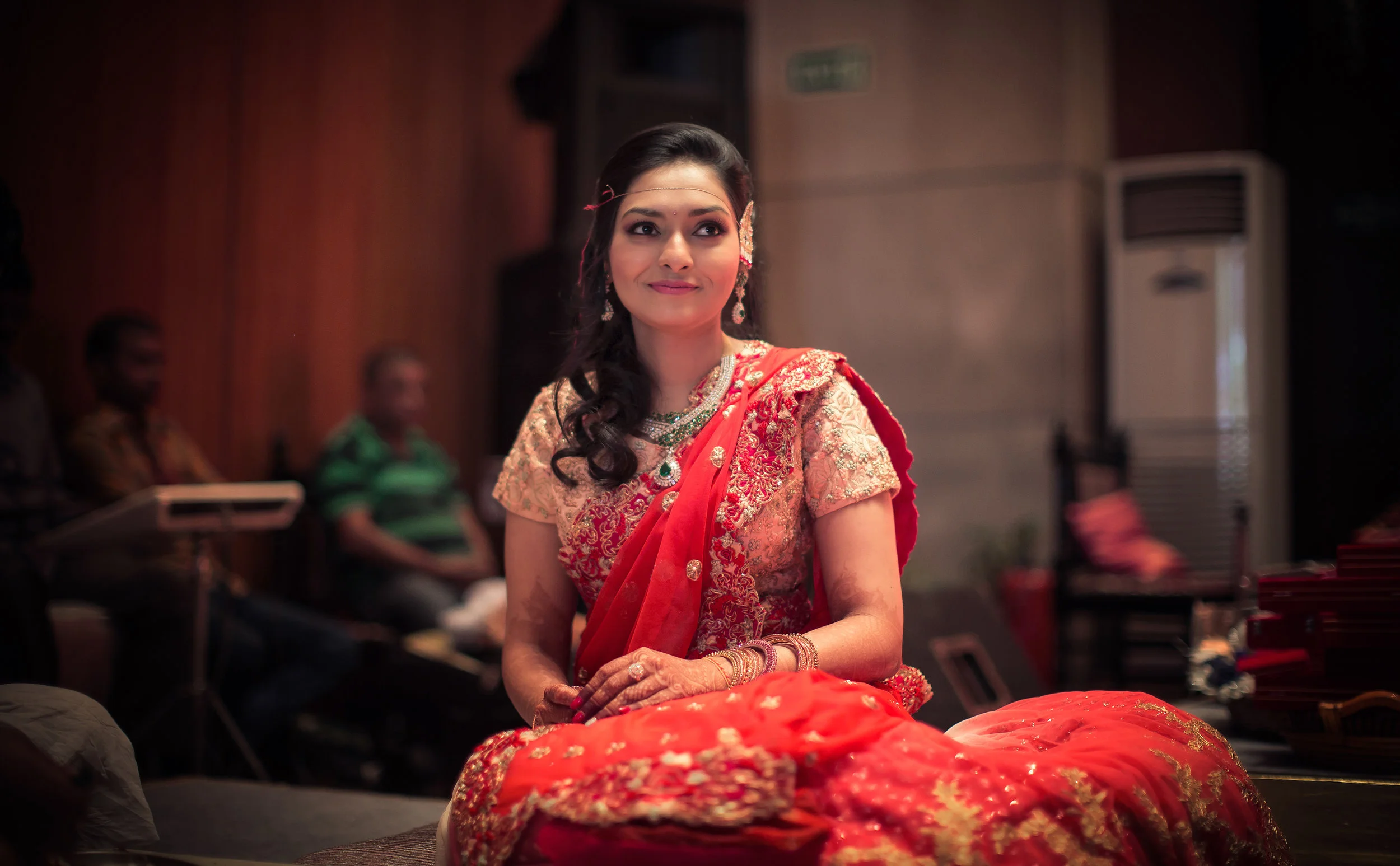 A woman in traditional Indian attire, wearing a red and gold saree with jewelry, sitting indoors with a soft smile.