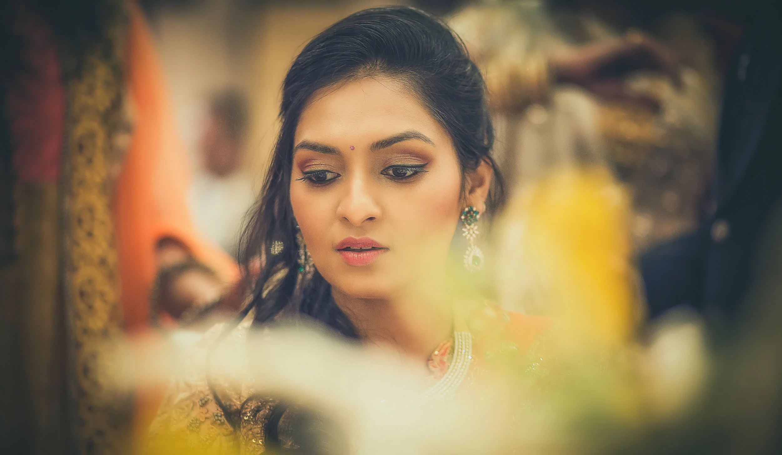 A woman dressed in traditional Indian attire with jewelry, looking down thoughtfully, with blurred figures and decorative elements in the background.