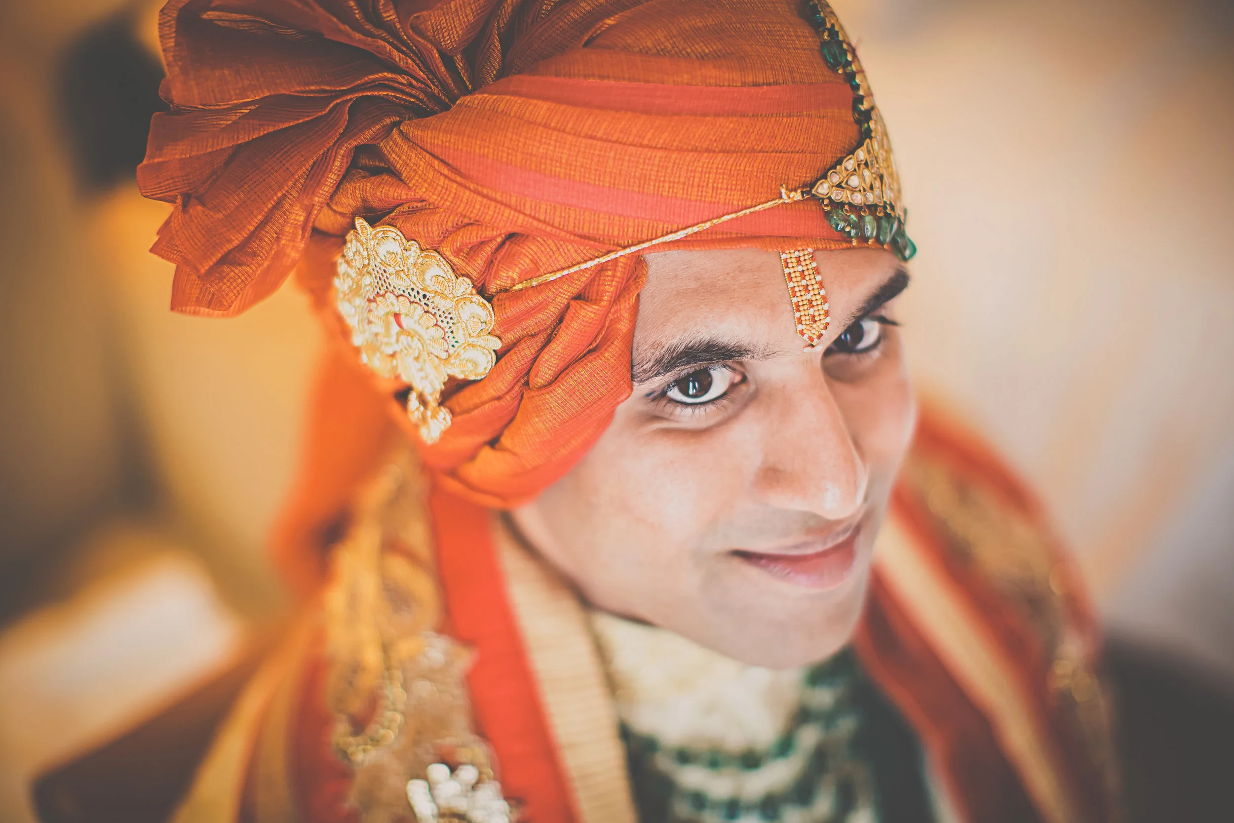 A man dressed in traditional Indian wedding attire with an orange turban adorned with gold embellishments and jewelry, smiling softly.