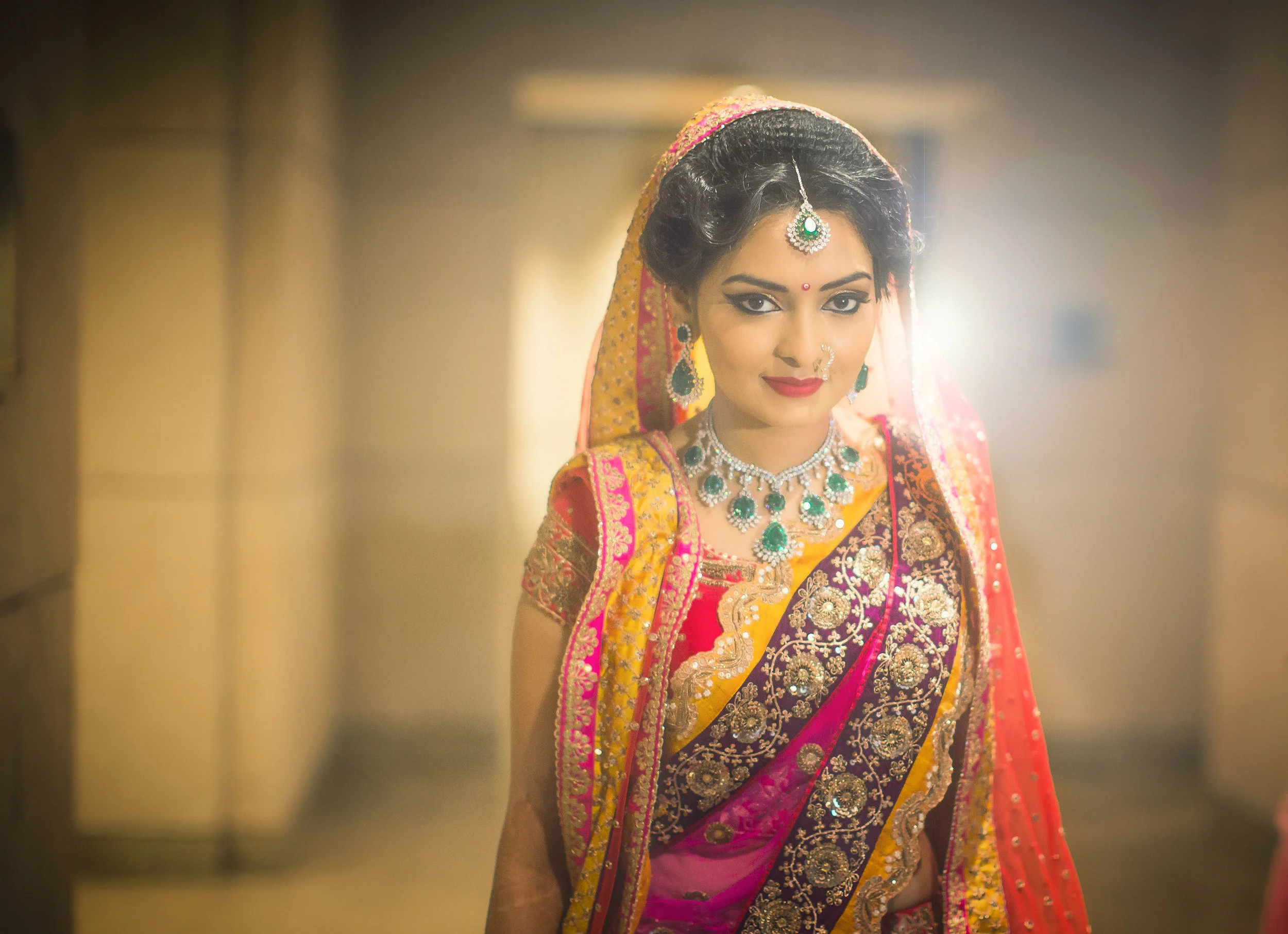 A woman wearing a traditional Indian saree with intricate embroidery, jewelry, and makeup, standing in a warmly lit indoor setting.