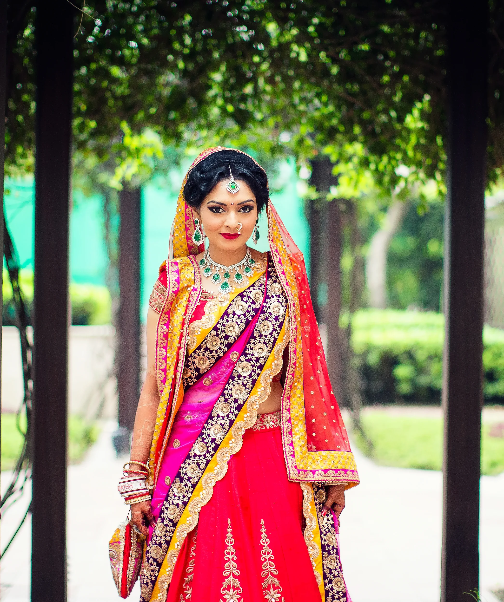 A woman dressed in traditional Indian bridal attire with vibrant pink, purple, and gold embroidery, wearing jewelry, standing outdoors under trees with a blurred green background.
