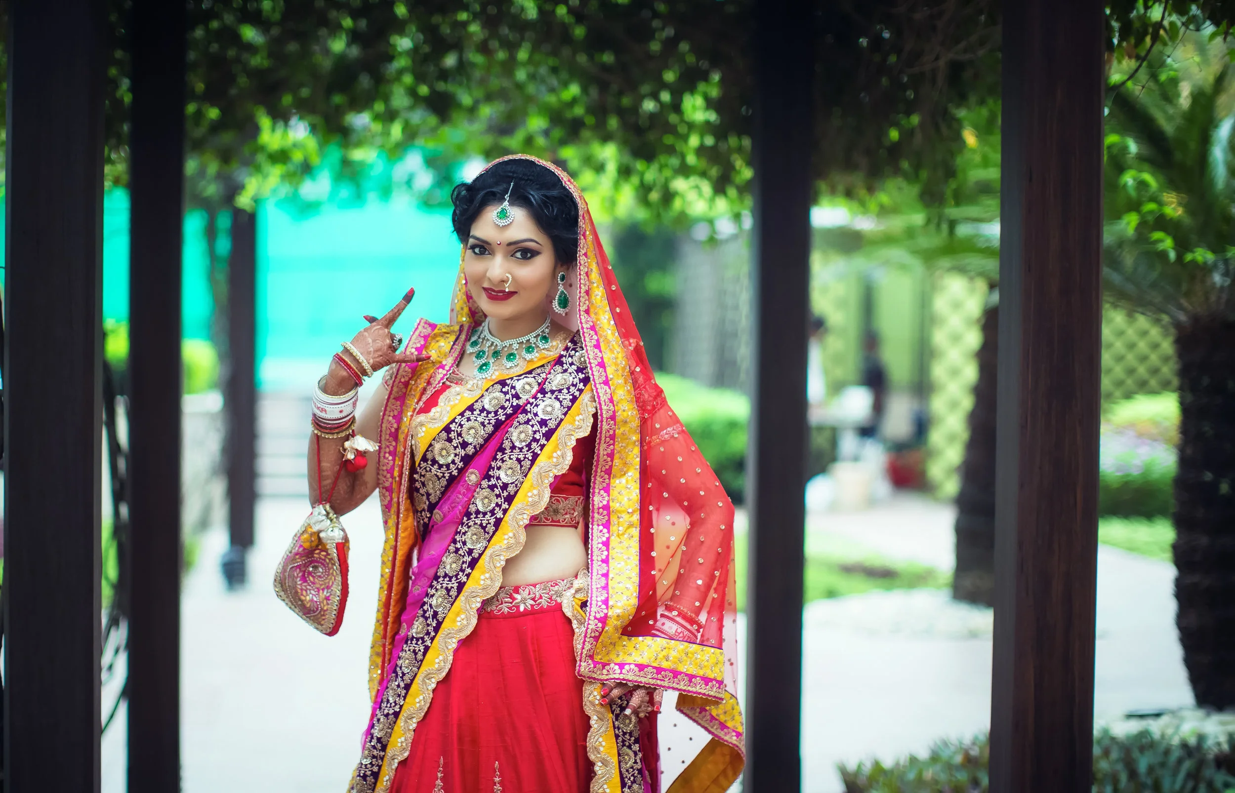 A woman dressed in traditional Indian attire, wearing a red, purple, and gold saree with jewelry, standing outdoors amidst greenery.
