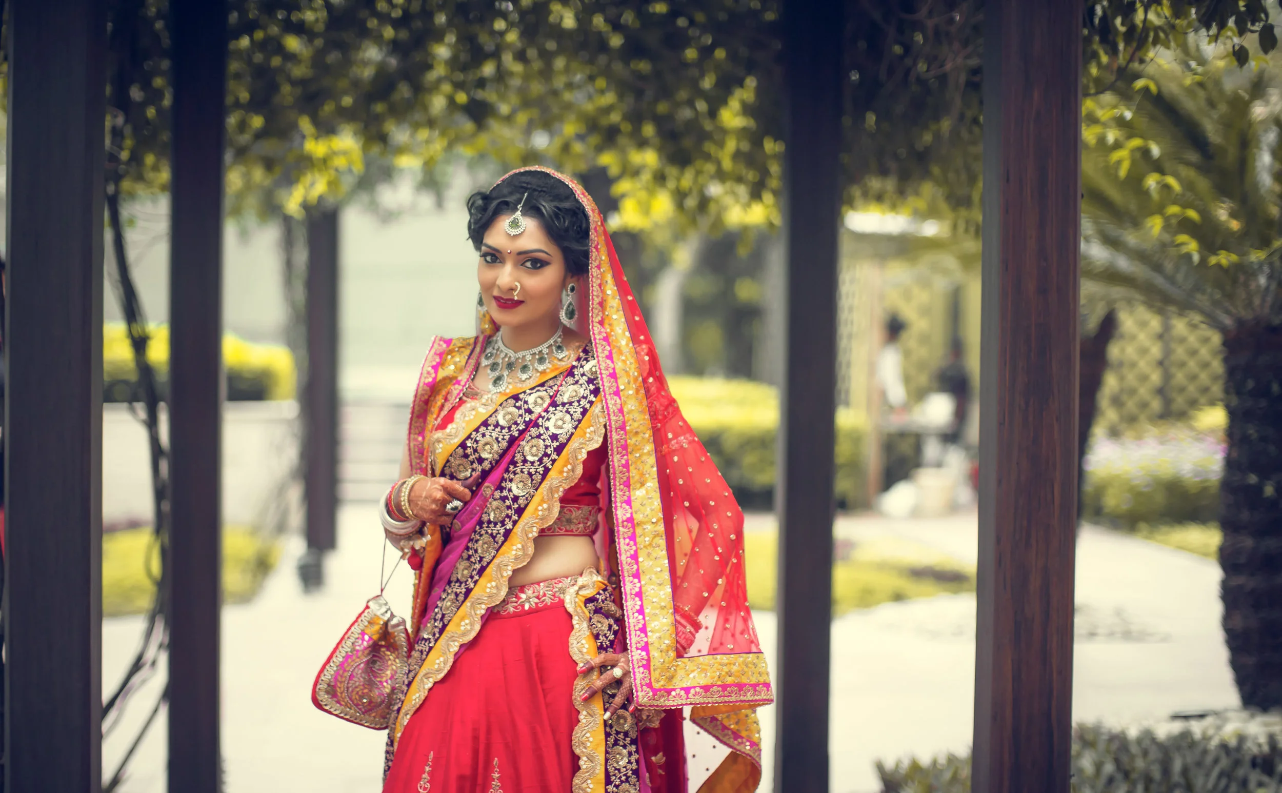 A woman dressed in traditional Indian bridal attire, wearing a red and gold saree with intricate jewelry, standing outdoors in a garden setting.