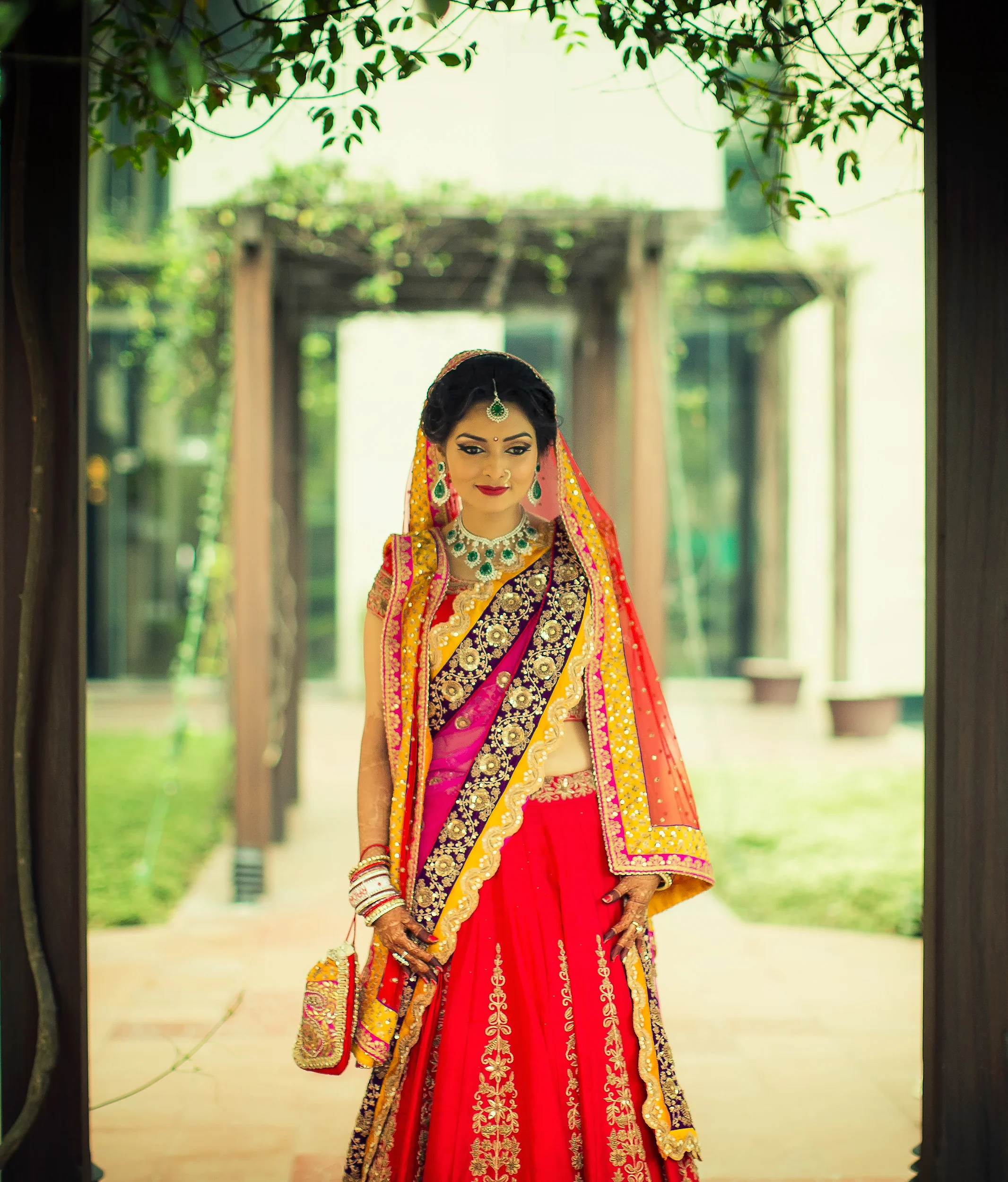 A woman dressed in traditional Indian wedding attire, standing outdoors and framed by a wooden doorway. She is wearing a vibrant red and gold embroidered lehenga, with jewelry including necklaces, earrings, and a maang tikka, and a colorful dupatta w