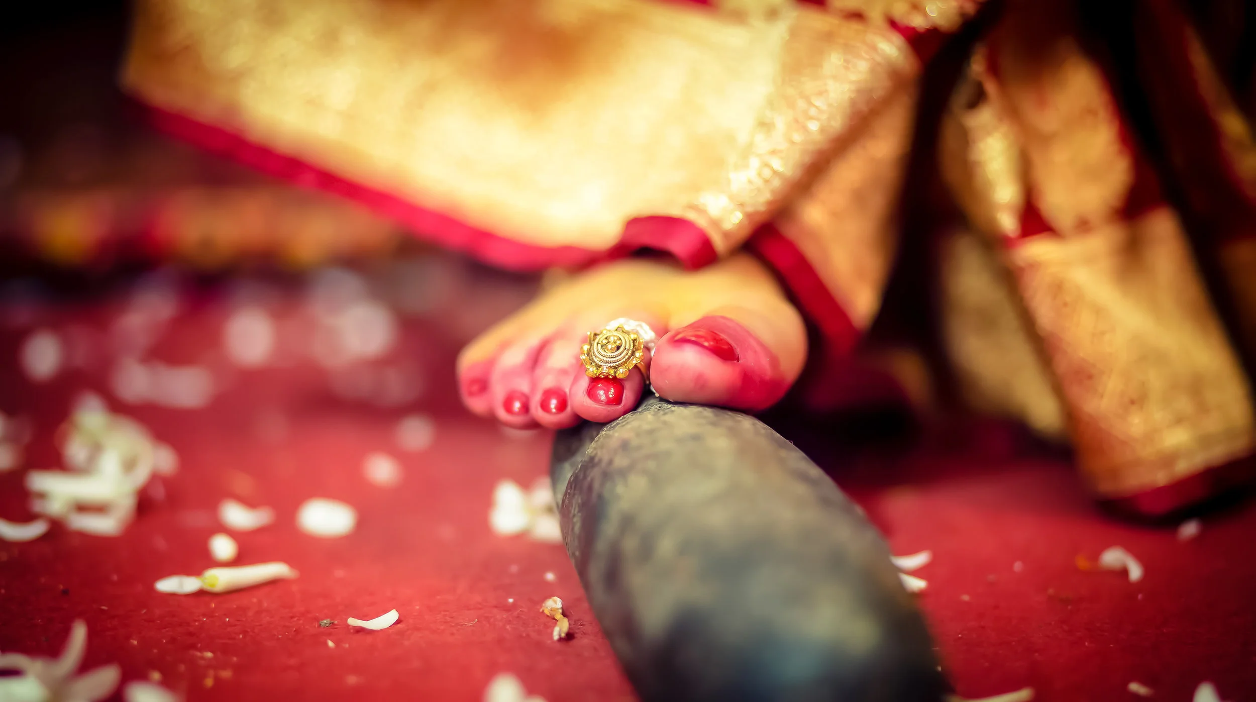 Close-up of a woman's foot with red toenail polish, wearing a traditional gold and pearl ring, as she places her foot on a ceremonial brass vessel during a wedding ritual, with flower petals scattered on a red carpet.