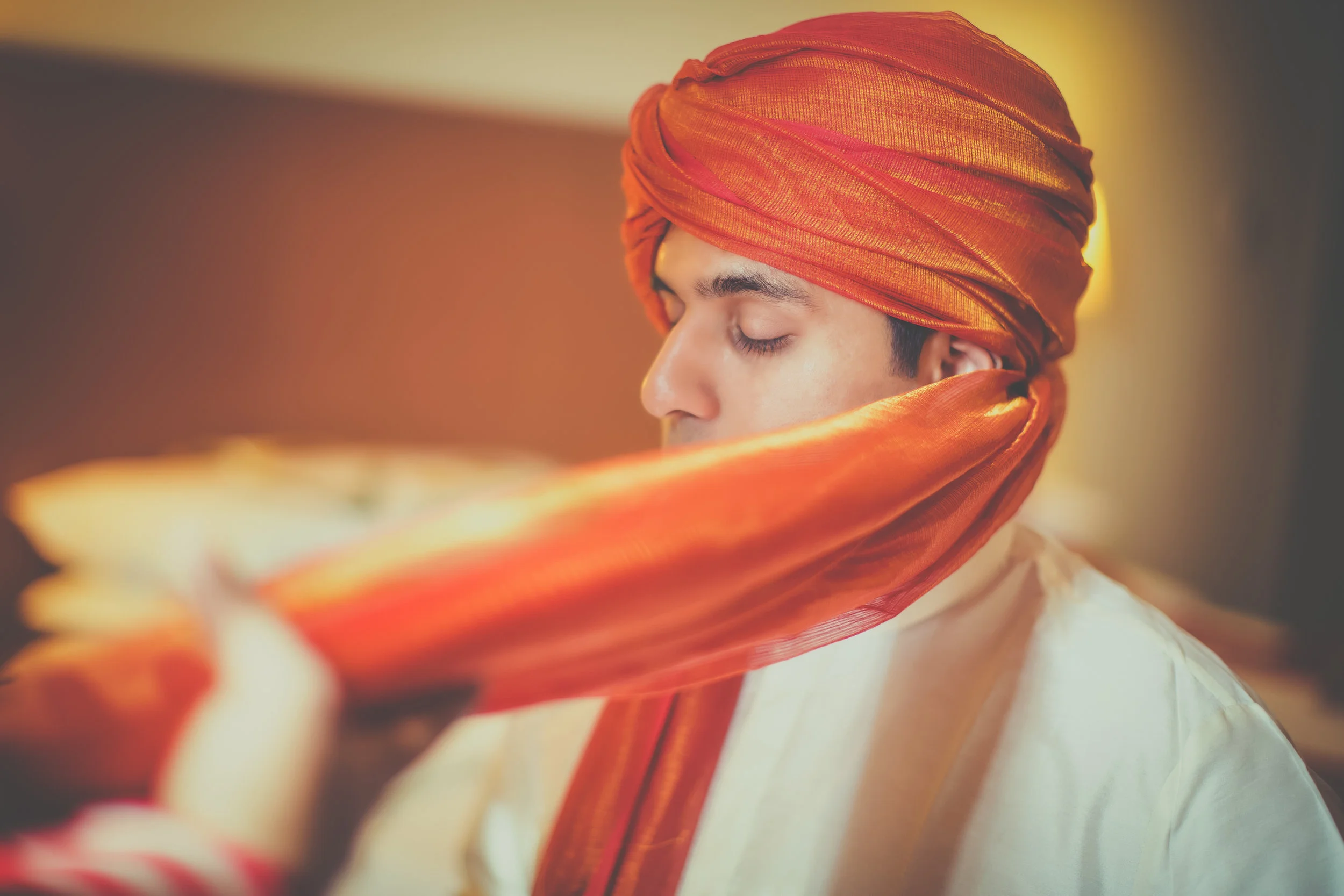 A man with his eyes closed is wearing a traditional orange turban and matching scarf, suggesting he is preparing for a cultural or religious event.