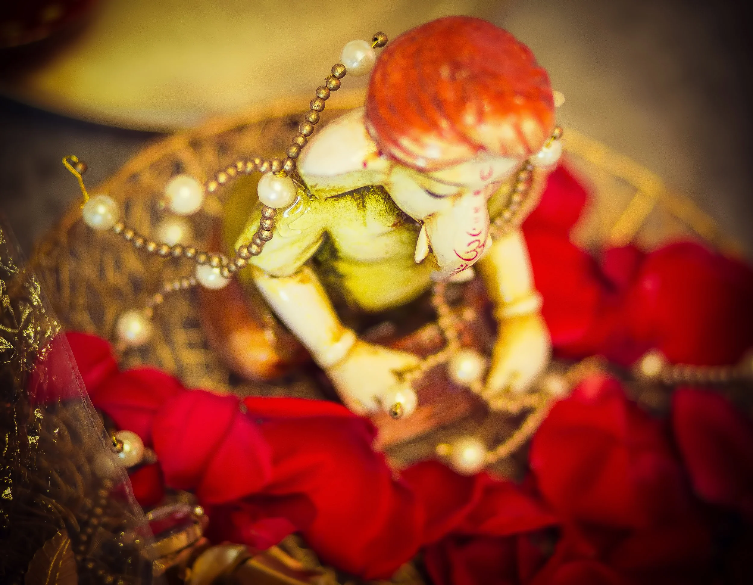 A small statue of Lord Ganesha sitting on a bed of red flower petals, adorned with pearl and gold jewelry, with a golden decorative background.