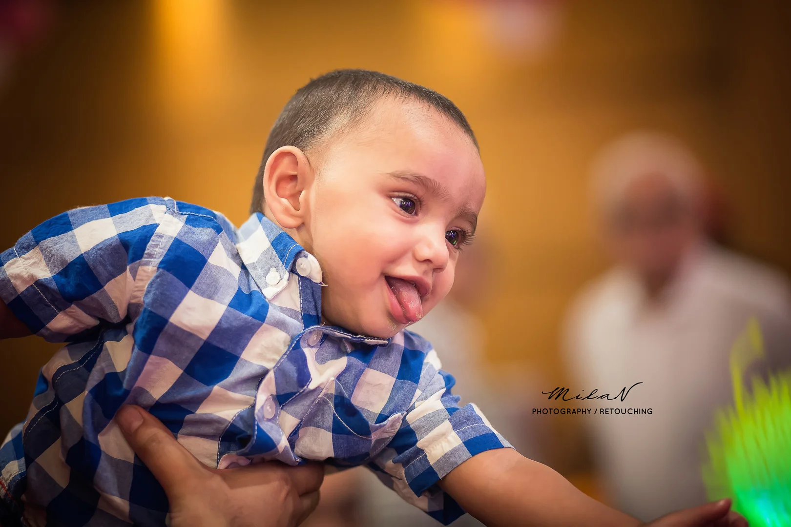 Close-up of a young boy in a blue and white checkered shirt, sticking out his tongue while being held by someone. The background is blurred with another child visible in the distance.