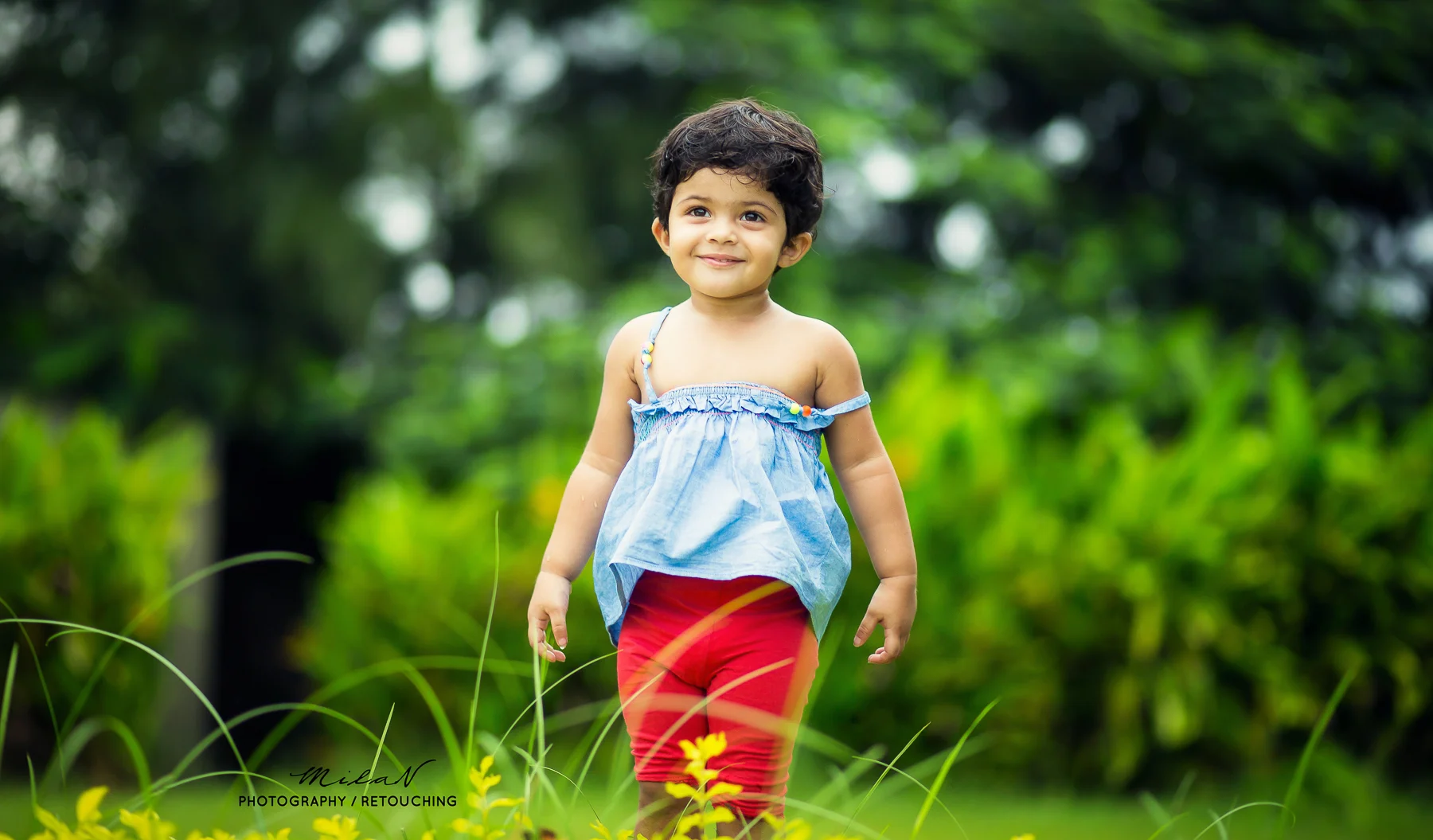 A young girl with dark hair and a smile, wearing a blue top and red pants, walking through a green outdoor area with plants and trees.