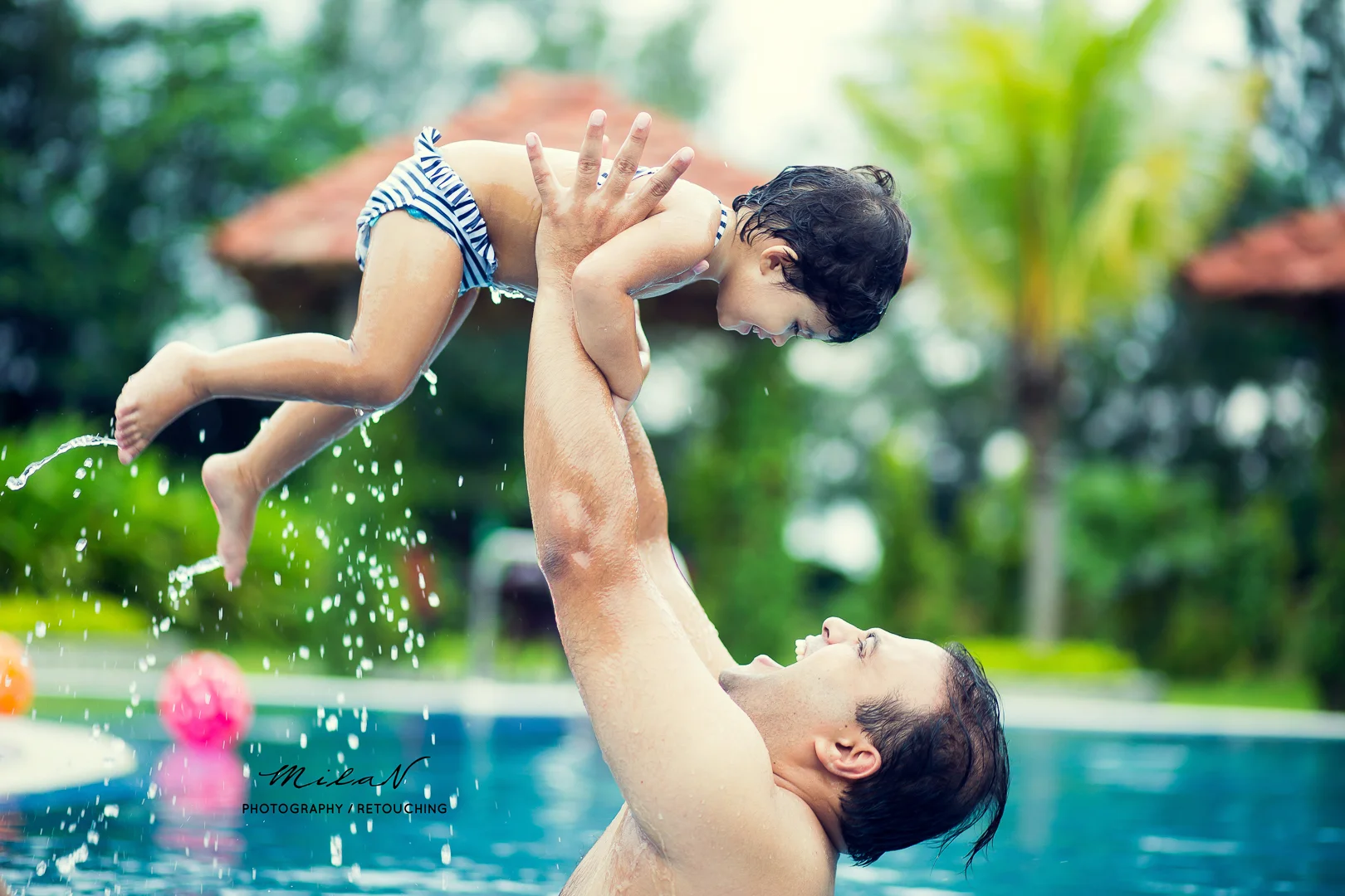 A woman lifting a young boy in a swimming pool, smiling and playing, with greenery and a gazebo in the background.