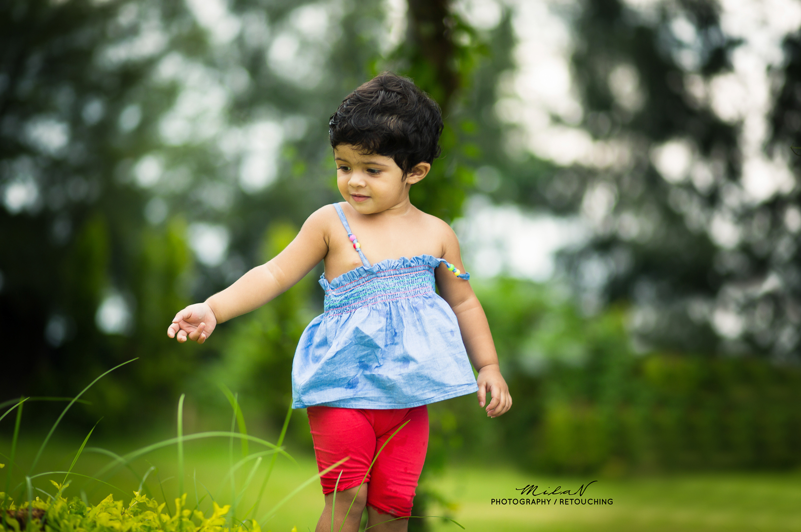Young girl exploring outdoors in a grassy area, wearing a blue dress and red shorts, surrounded by greenery.