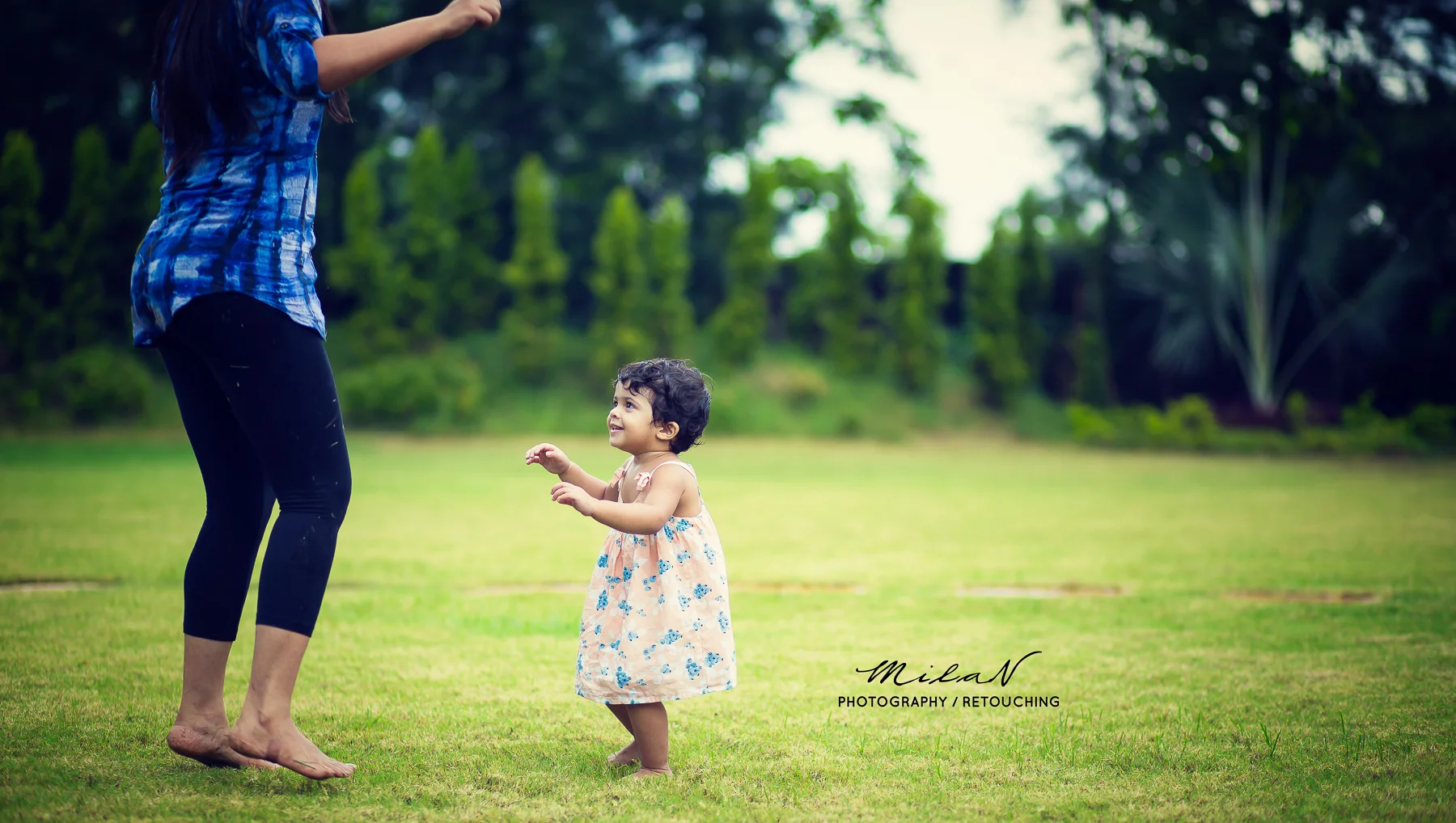 A young girl in a floral dress looking up and smiling at a woman in a blue plaid shirt and black pants, standing barefoot on grass in a park or garden.
