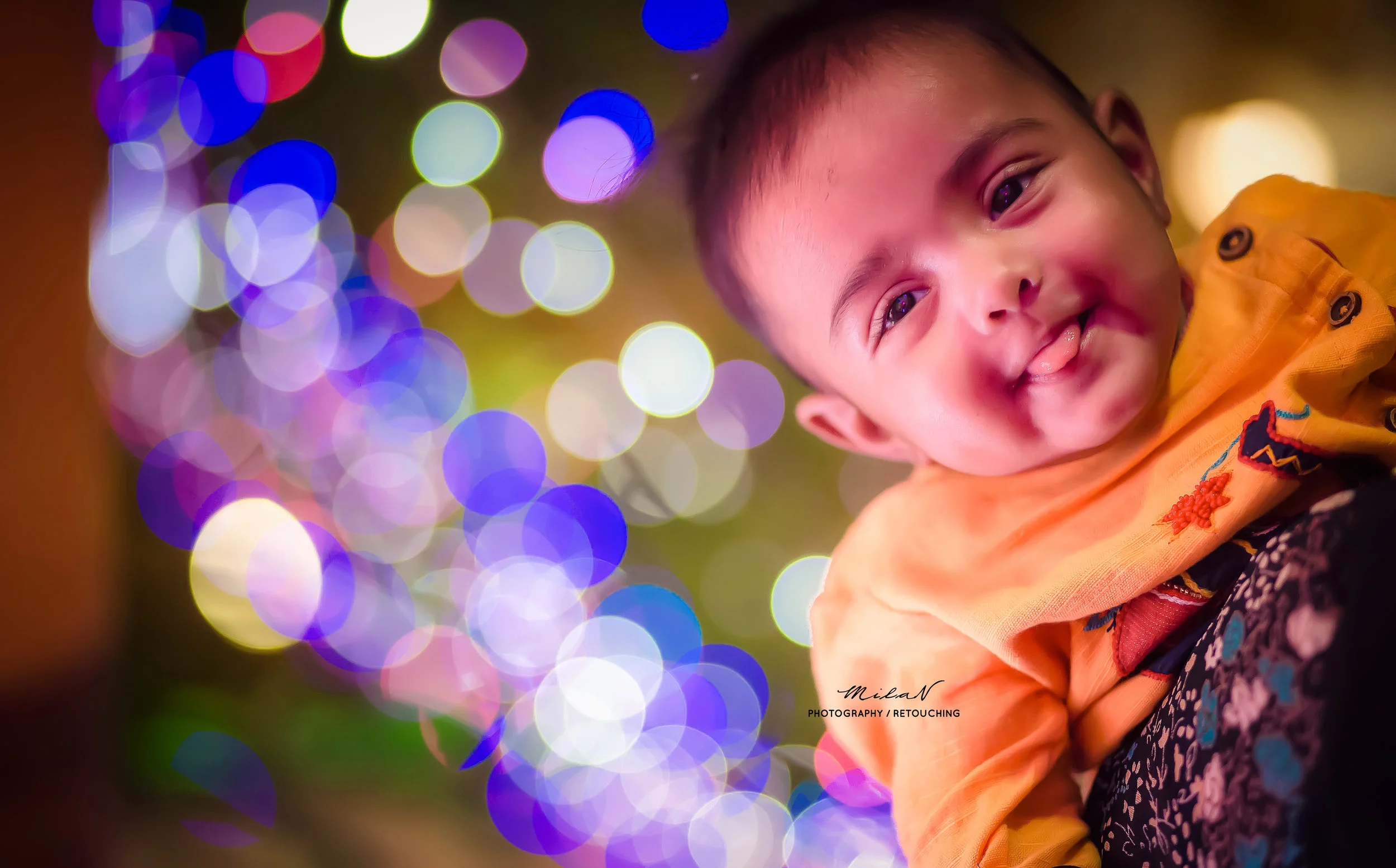 Smiling baby wearing orange clothing with colorful embroidery, with blurry multicolored lights in the background.