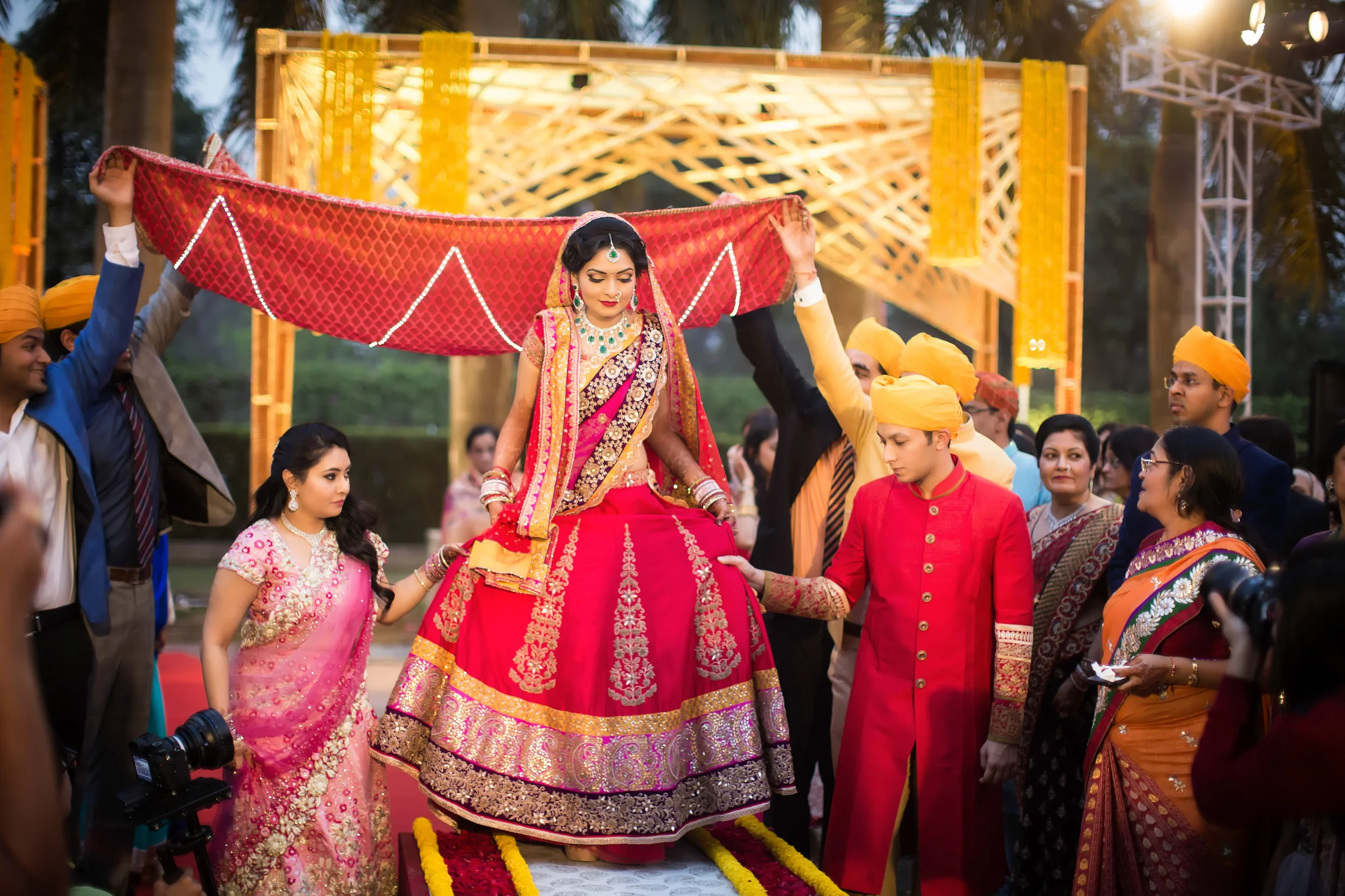 A traditional Indian wedding ceremony featuring a bride seated on a decorated platform, wearing a red and gold bridal outfit with jewelry, surrounded by family and friends dressed in colorful traditional attire. Men in yellow turbans are lifting a ca