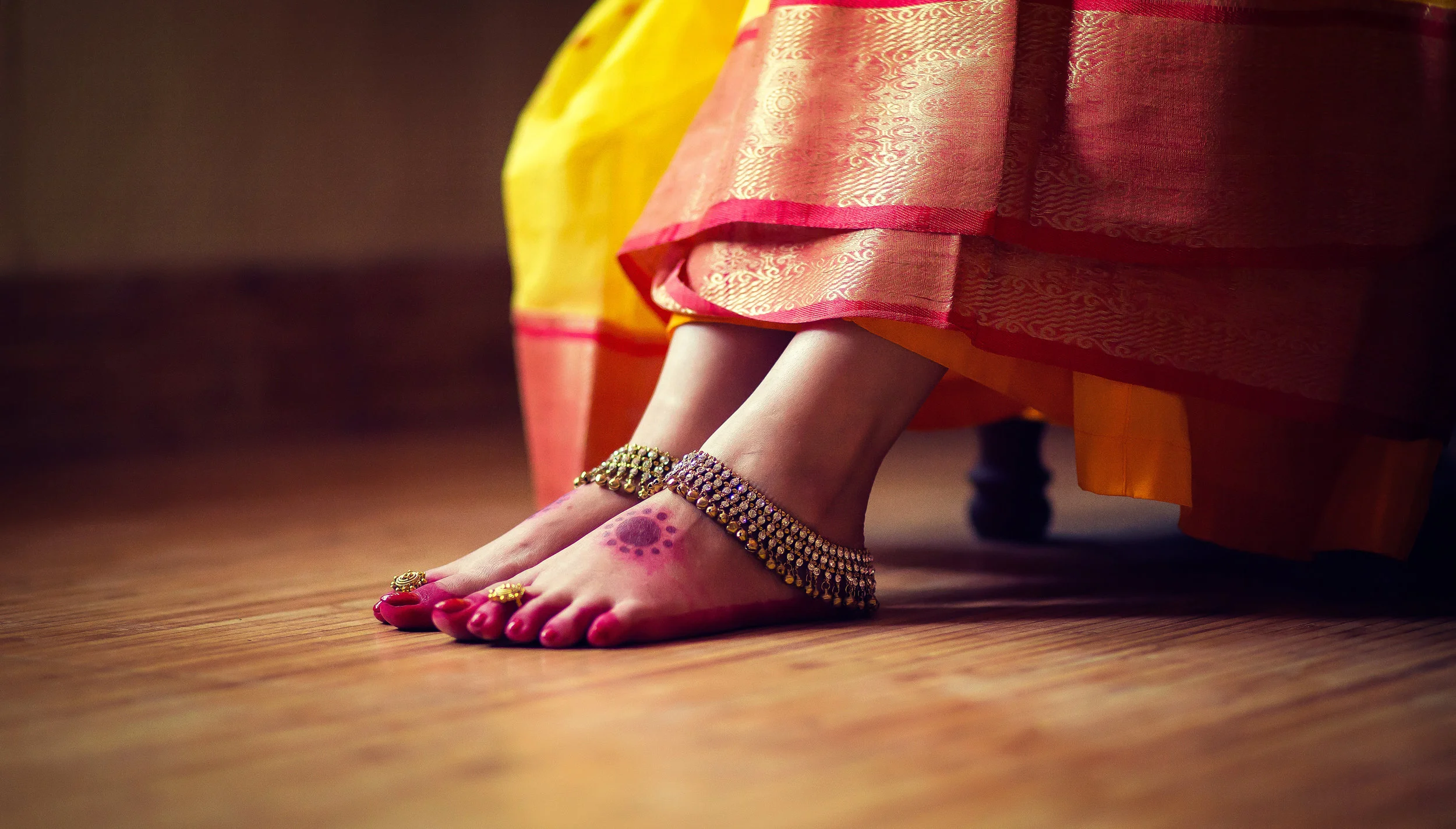 Close-up of a bride's feet in traditional Indian silk footwear, decorated with anklets and toe rings, with henna designs on her toes, as she sits on a wooden floor wearing a colorful saree.