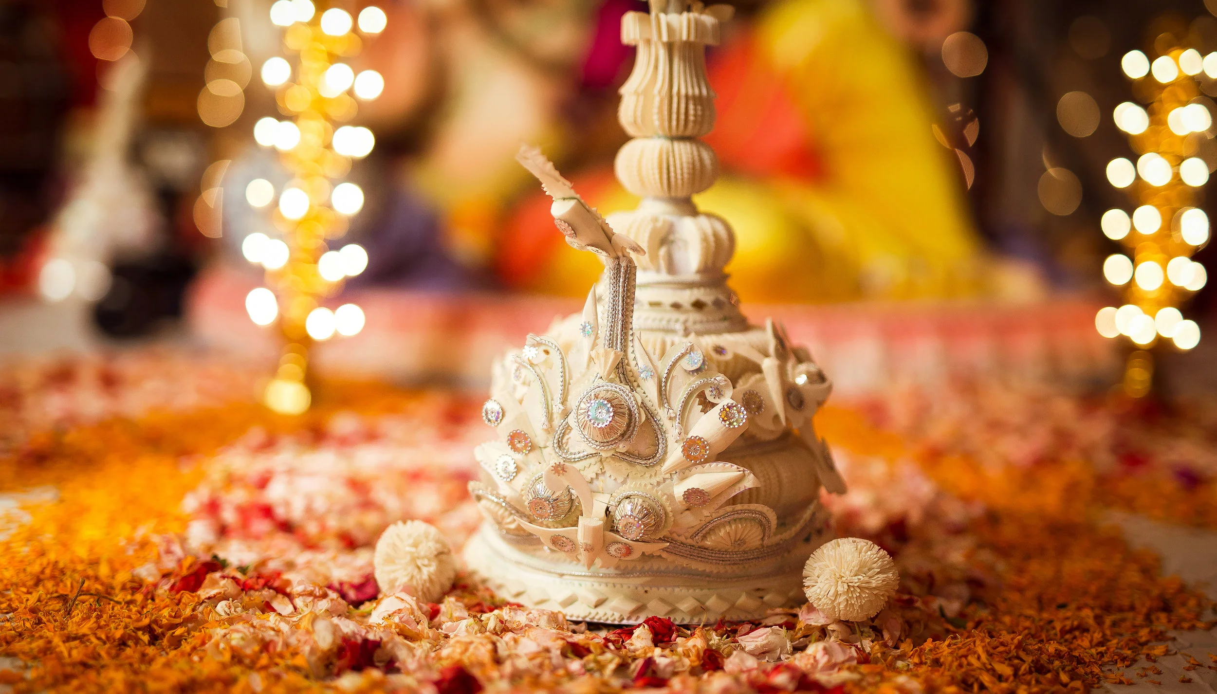 A decorative white and cream-colored Hindu Shiva Lingam adorned with jewelry, surrounded by orange and pink flower petals and two white flowers, with blurred warm lights in the background.