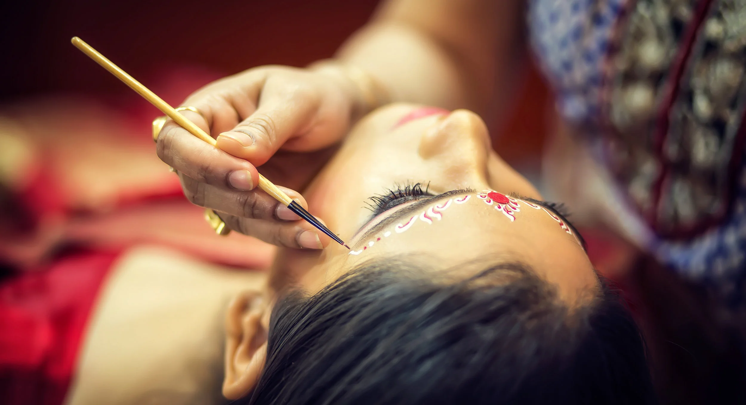 A woman lying down while an artist using a thin brush to paint intricate designs and patterns on her face, including makeup around her eyes and forehead.