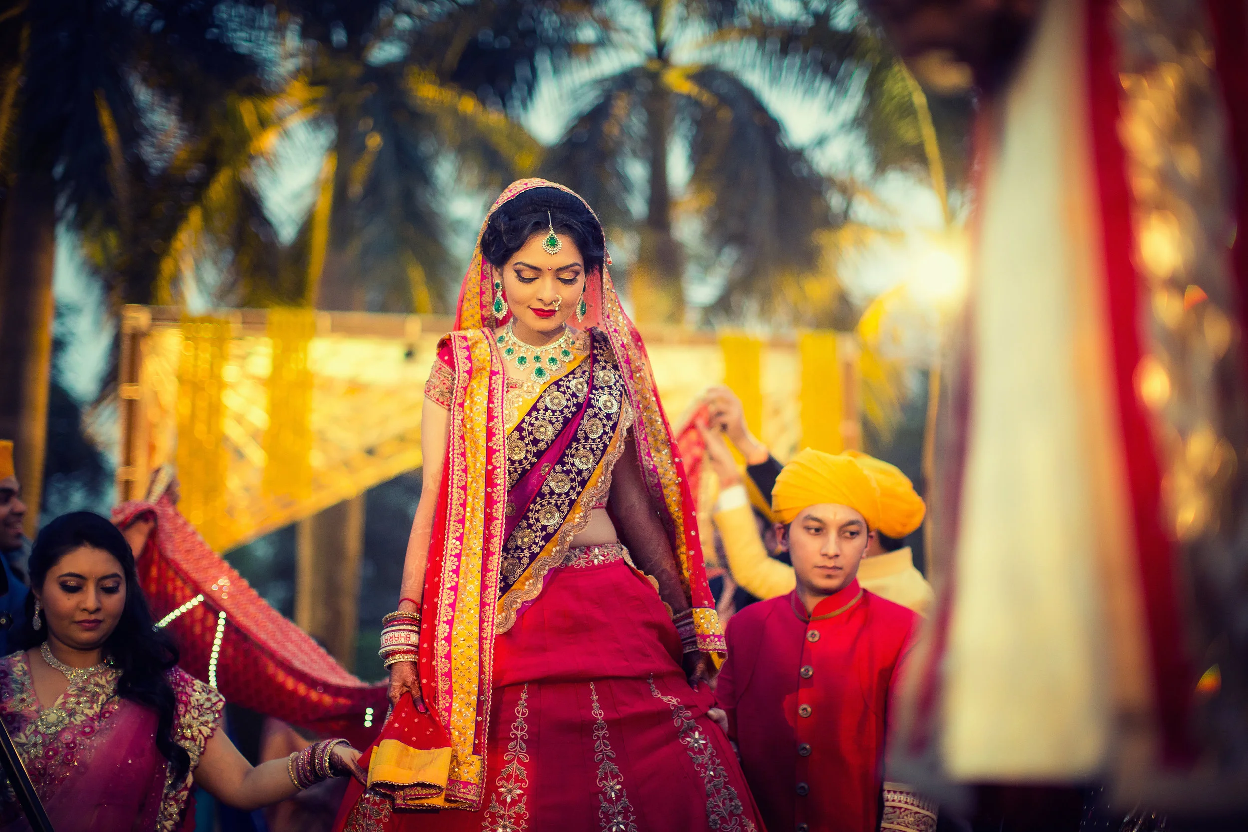A bride dressed in traditional Indian wedding attire, wearing a pink and gold Lehenga, with intricate jewelry, walking outdoors during sunset surrounded by people and palm trees.