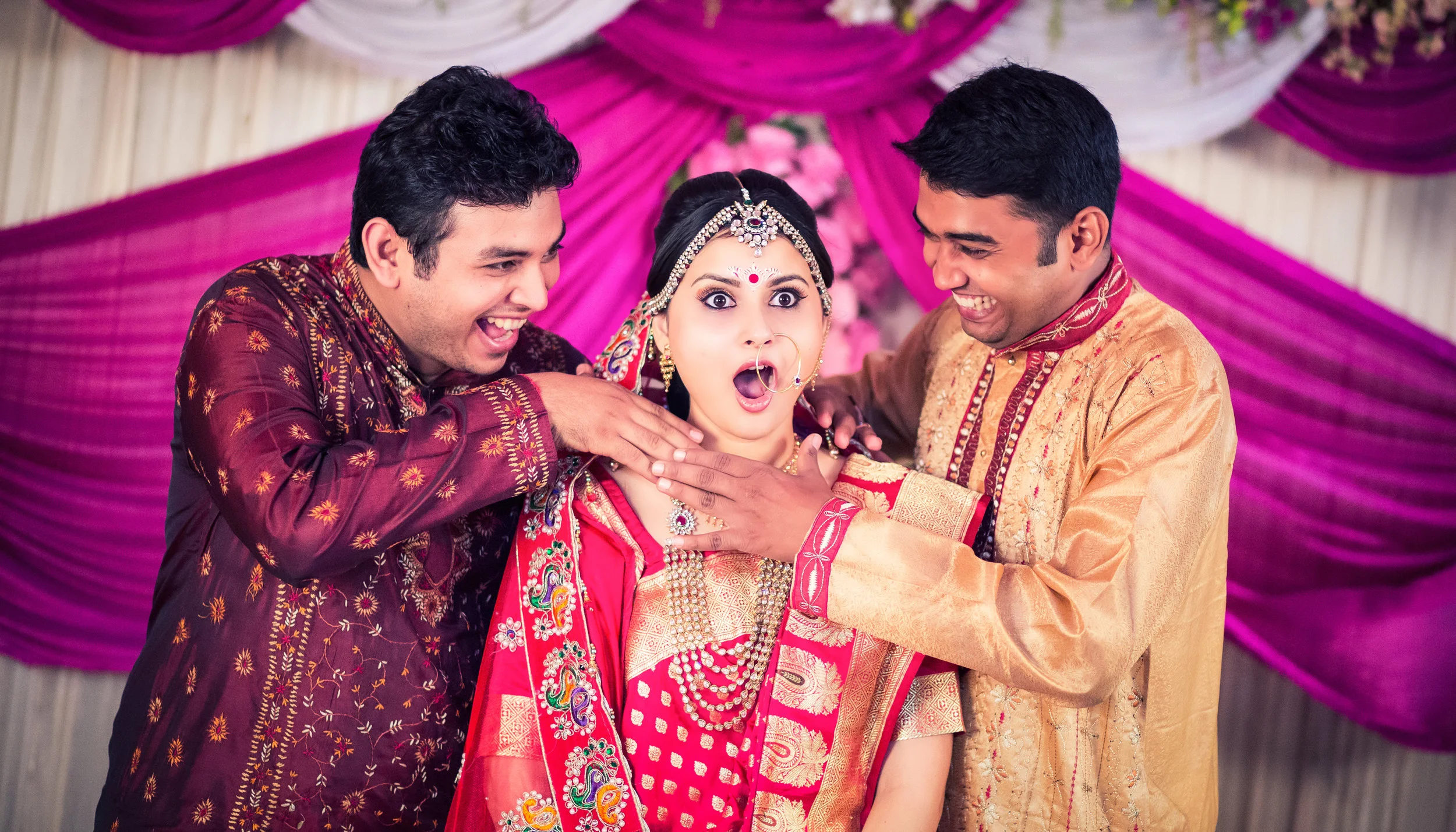 Three people in traditional Indian attire at a wedding, with the woman in the center wearing jewelry and a bright pink and gold dress, showing a surprised expression, surrounded by two smiling men.