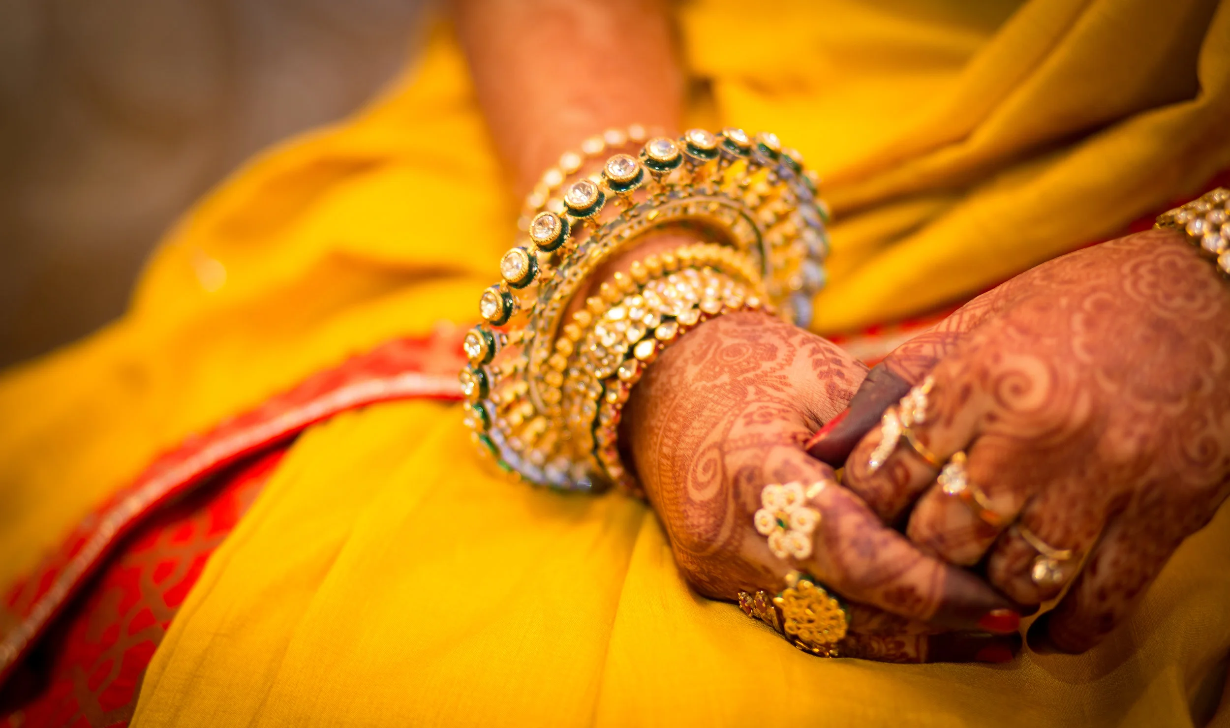 Close-up of a person's hand decorated with henna and wearing multiple gold and gemstone bangles, dressed in a yellow and red traditional outfit.
