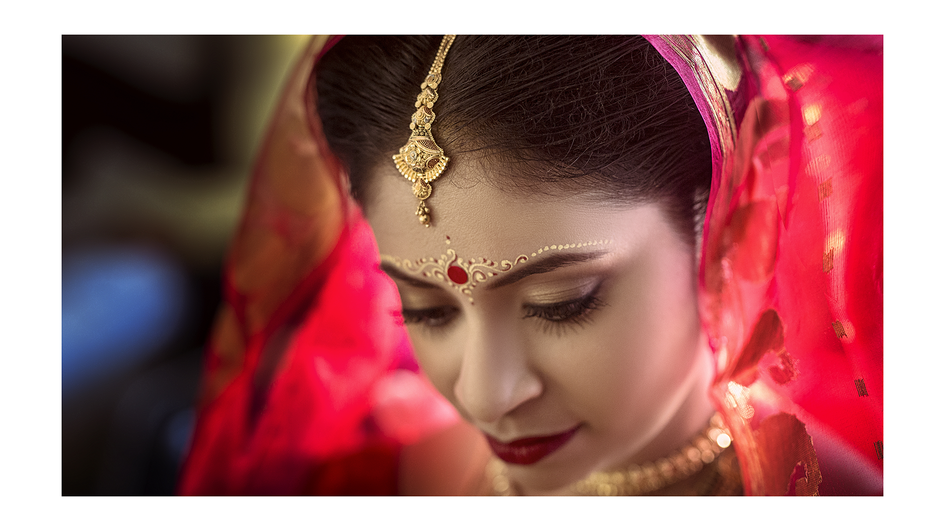 Close-up of a woman in traditional Indian attire, wearing a red saree with gold jewelry, including a headpiece and face decoration, with her eyes closed.