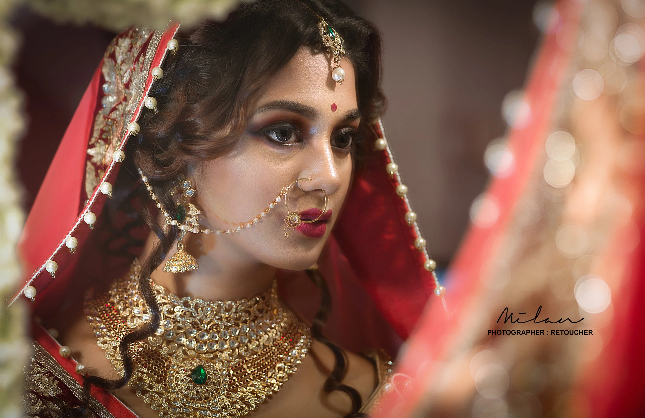 Close-up of an Indian bride wearing traditional jewelry including a nose ring, earrings, necklace, and headpiece, with red and gold attire and makeup.