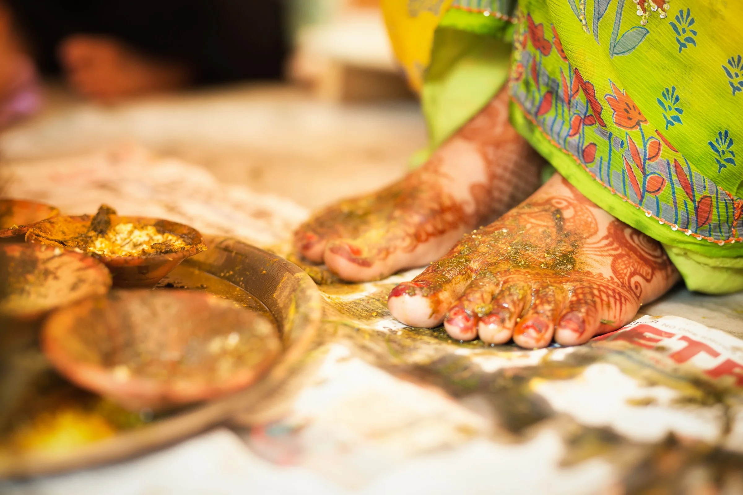 Close-up of a woman's feet covered in henna designs, next to traditional Indian bowls of colorful spices or pastes, on a white surface, with part of her vibrant green and colorful embroidered clothing visible.