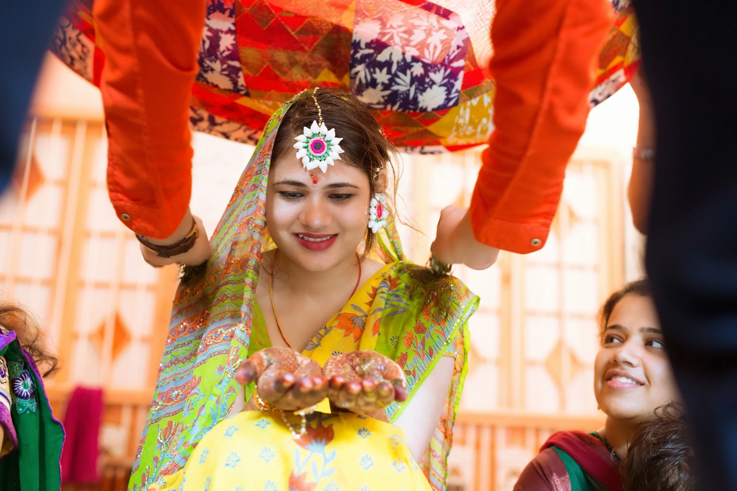 Young woman dressed in traditional colorful Indian saree, smiling and holding a small clay pot with her hands, participating in a cultural or religious ceremony. She is adorned with festive jewelry and body art, with a bigger person above pouring wat