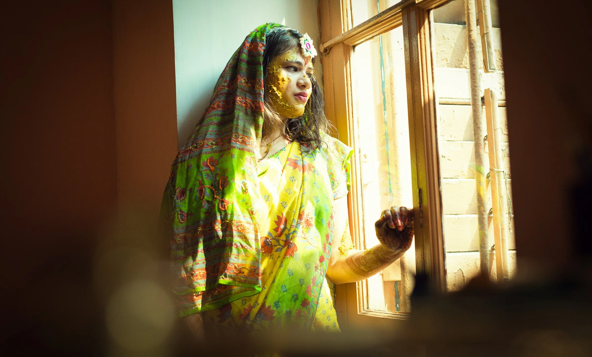 A woman dressed in traditional Indian attire, standing by a window with sunlight shining on her face, with yellow powder on her face and hands, and a floral headband.