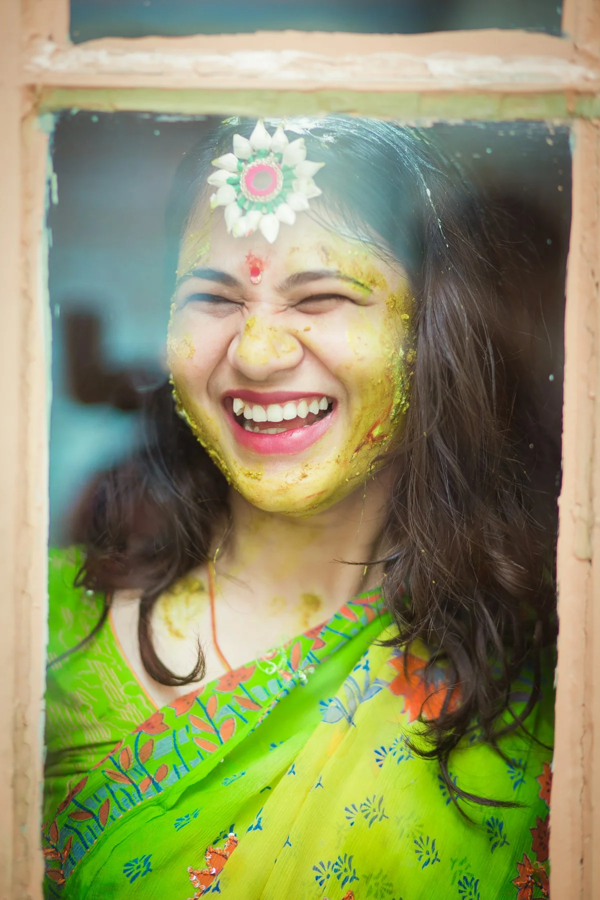 A woman smiling and laughing, covered in yellow and green colored powder, celebrating Holi festival, wearing a green traditional saree and a decorative headpiece.