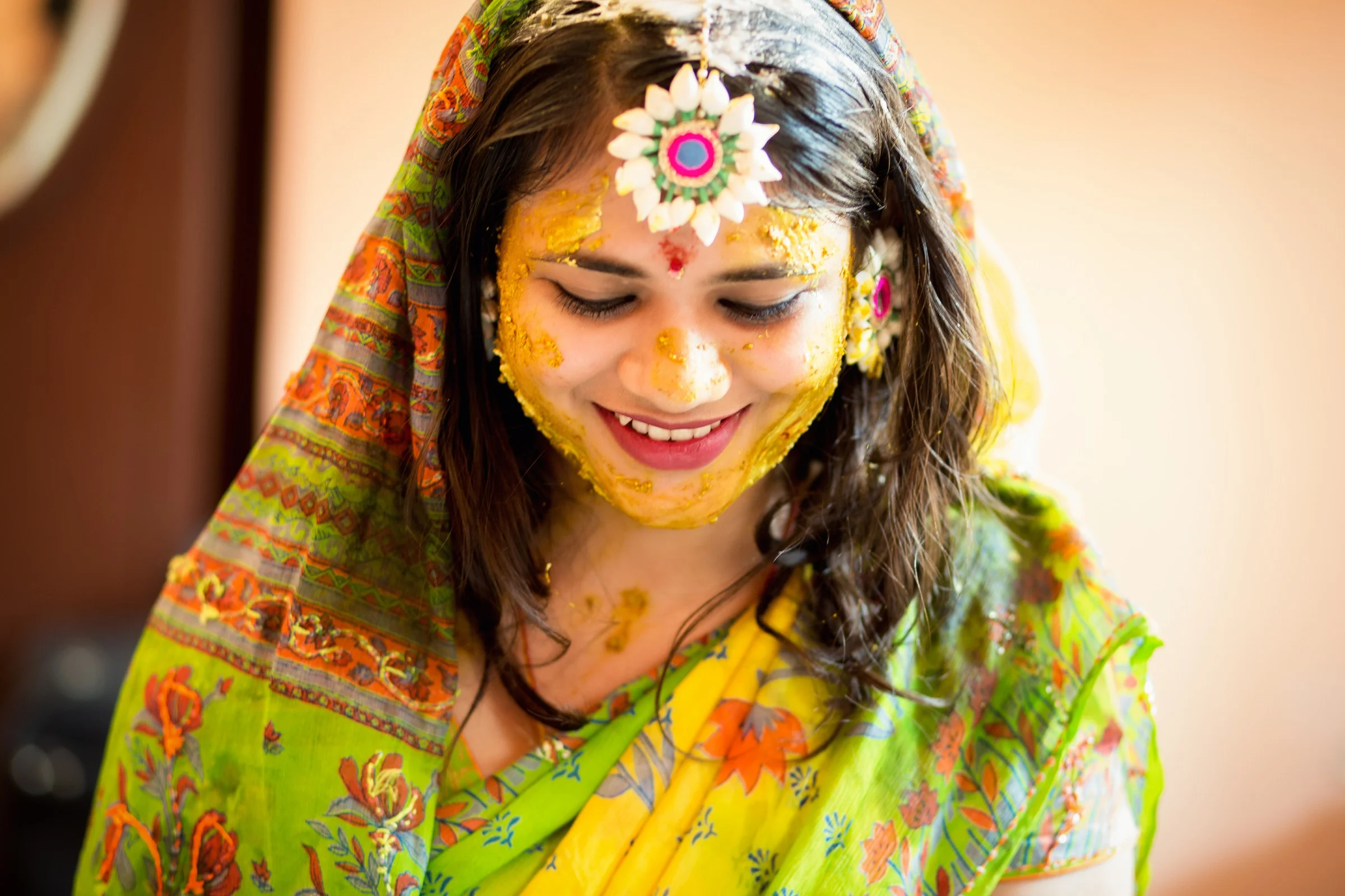 Young woman celebrating Holi, covered in yellow and orange powdered colors, wearing a colorful traditional sari and jewelry, smiling with her eyes closed.