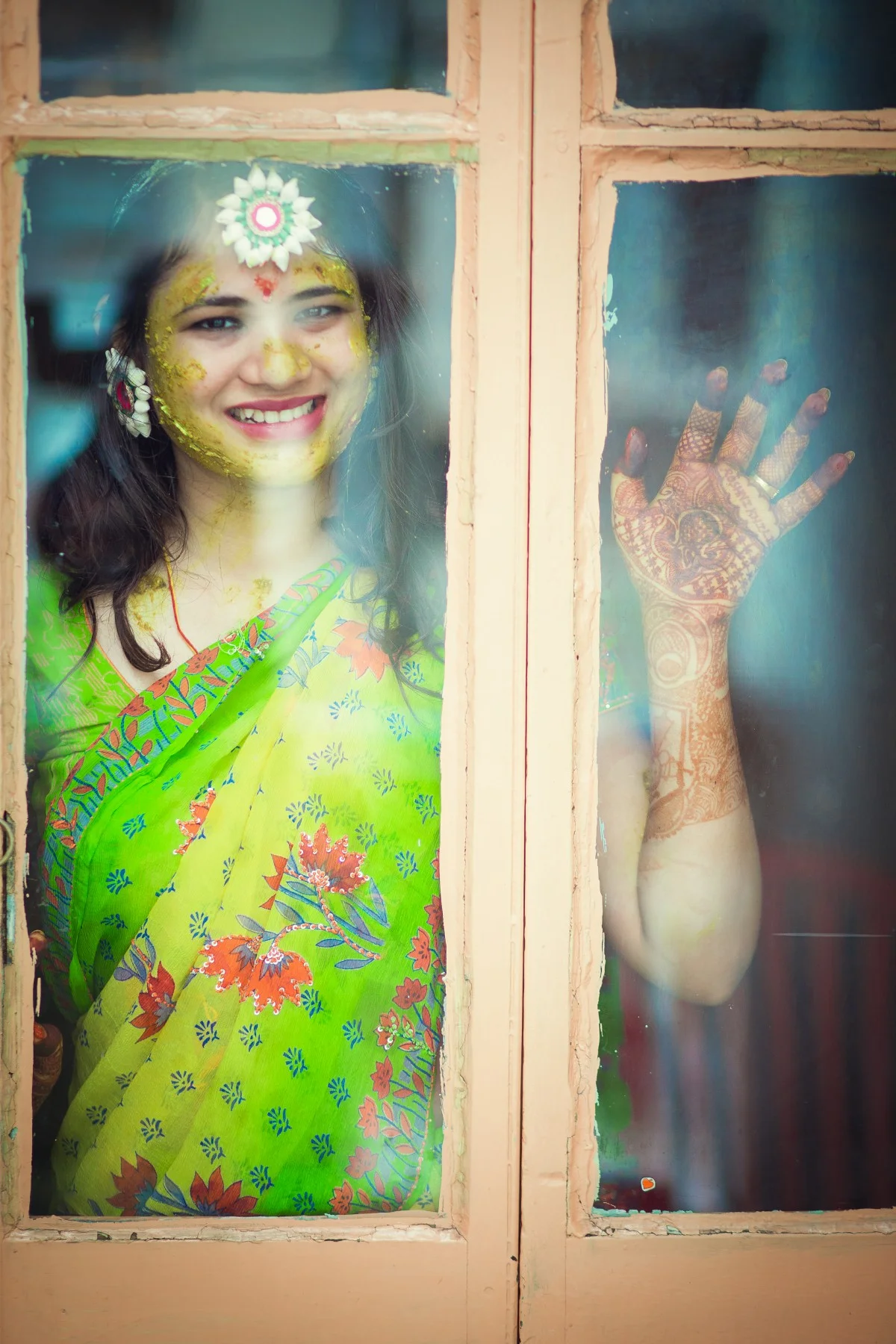 A woman in a green sari standing behind a window with a cheerful smile, decorated with yellow powder on her face, wearing jewelry, with a henna design on her arm and hand pressed against the glass.