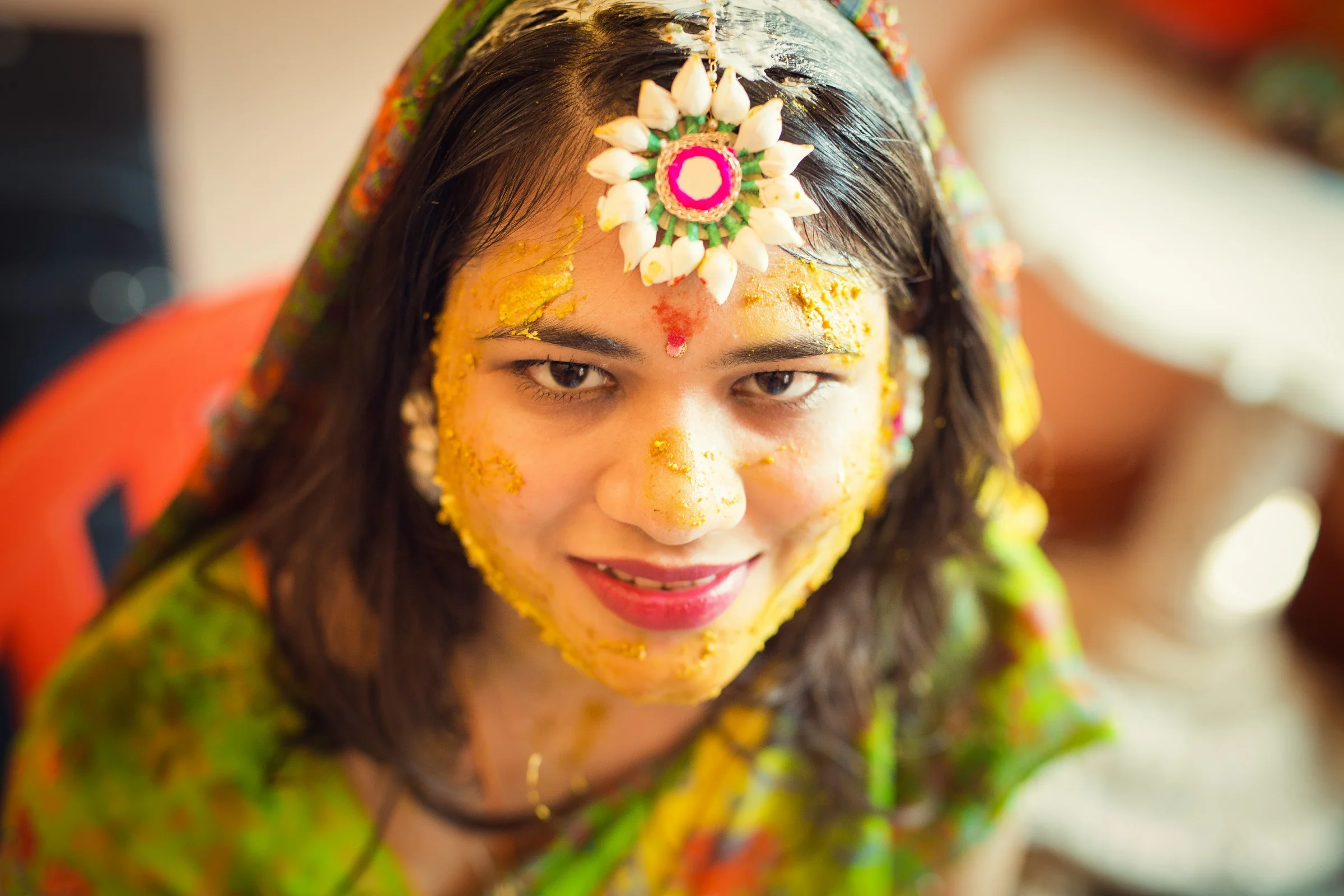 Young woman with a flower ornament headpiece, yellow paste on her face, and traditional attire during a cultural celebration.