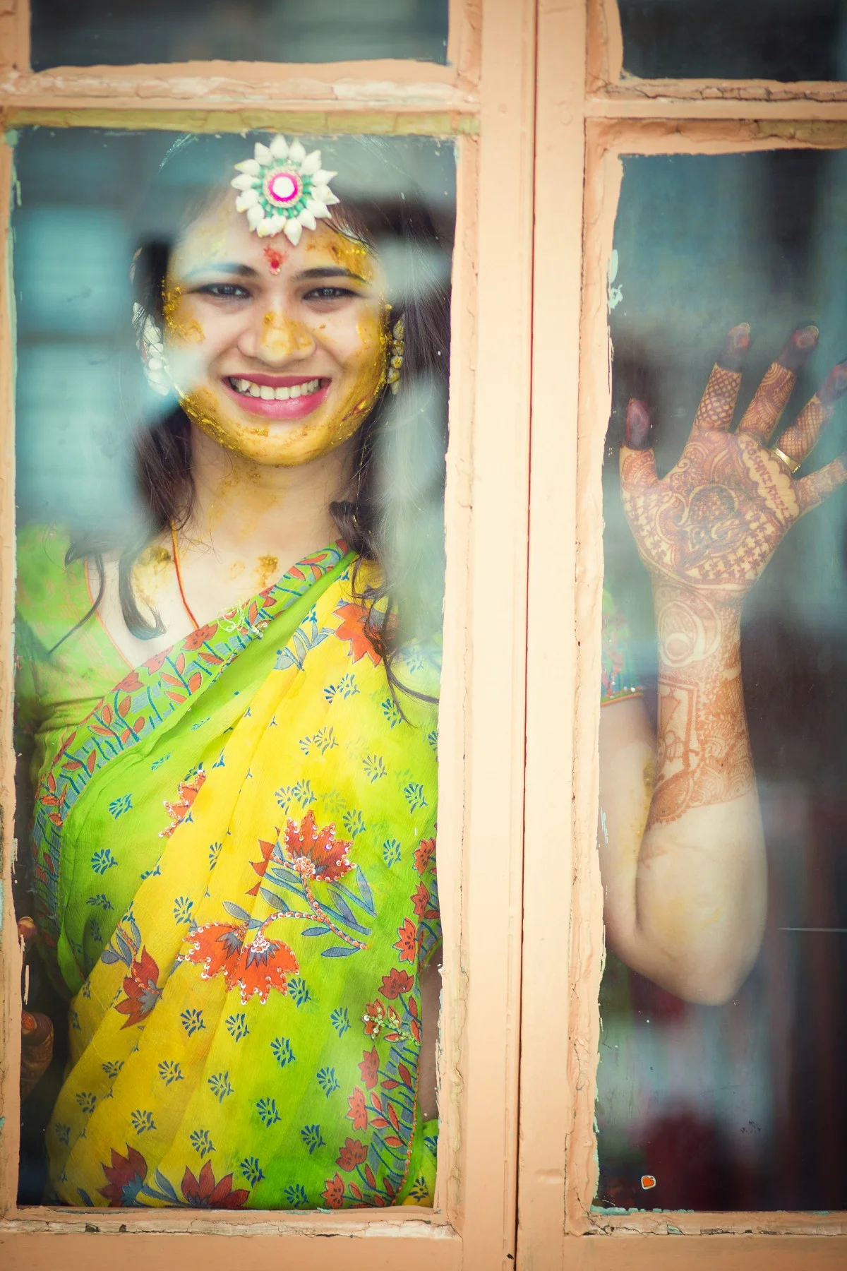 A woman dressed in a colorful sari, smiling and looking through a glass window, with her face and hand decorated for a celebration.