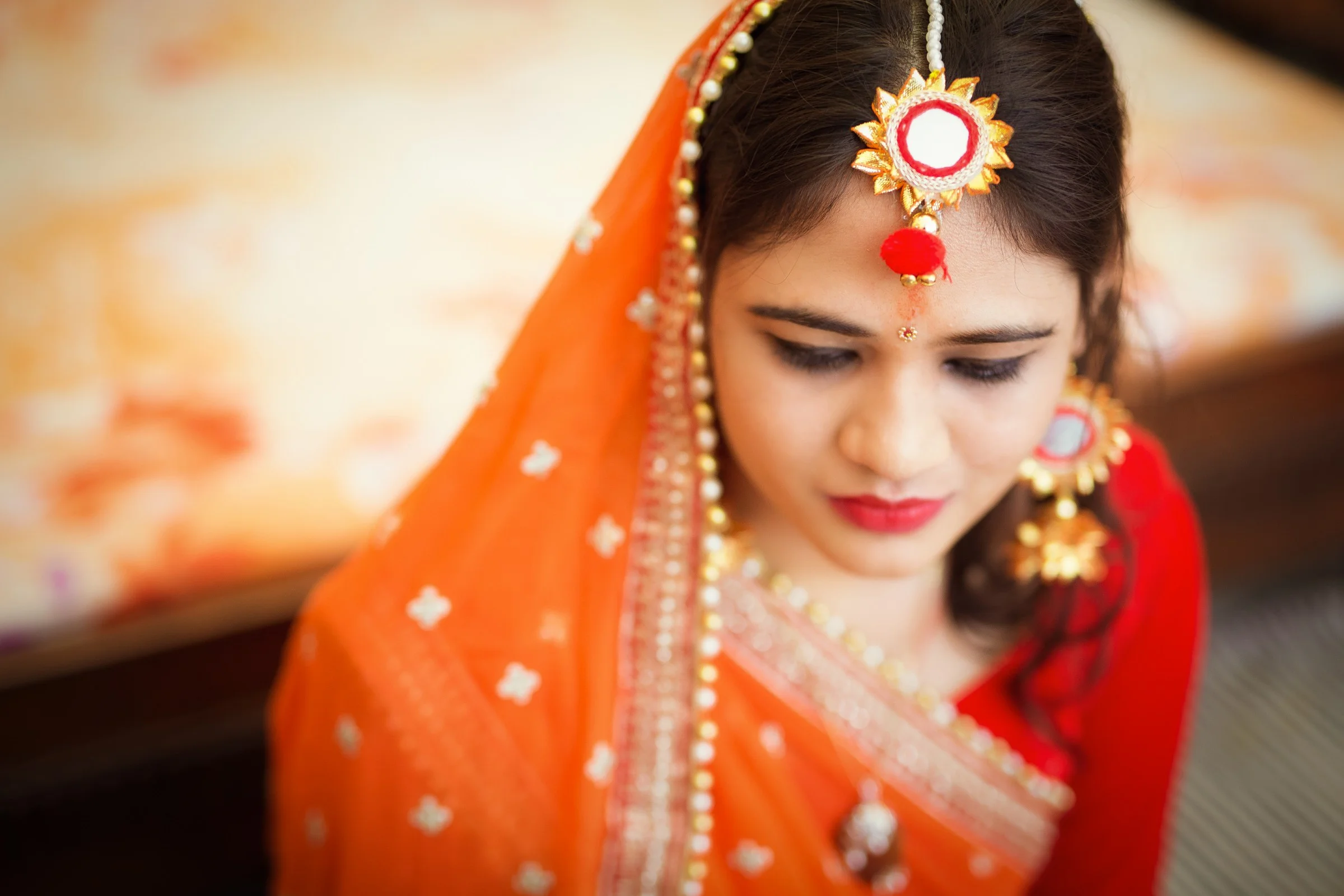 Young woman in traditional Indian attire, dressed in a bright orange saree with gold and white embellishments, wearing jewelry including earrings, a necklace, and a headpiece, with a red bindi on her forehead, looking downward.