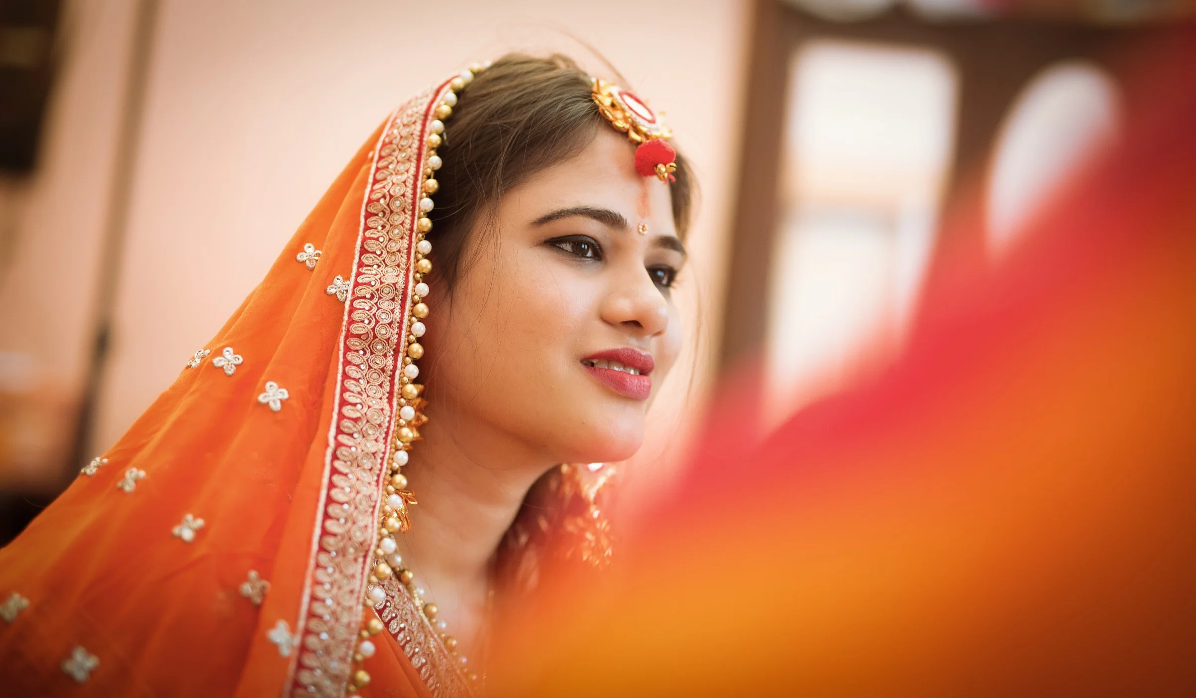 A woman dressed in traditional Indian attire, wearing an orange saree with intricate embroidery, jewelry, and a red bindi, smiling softly.