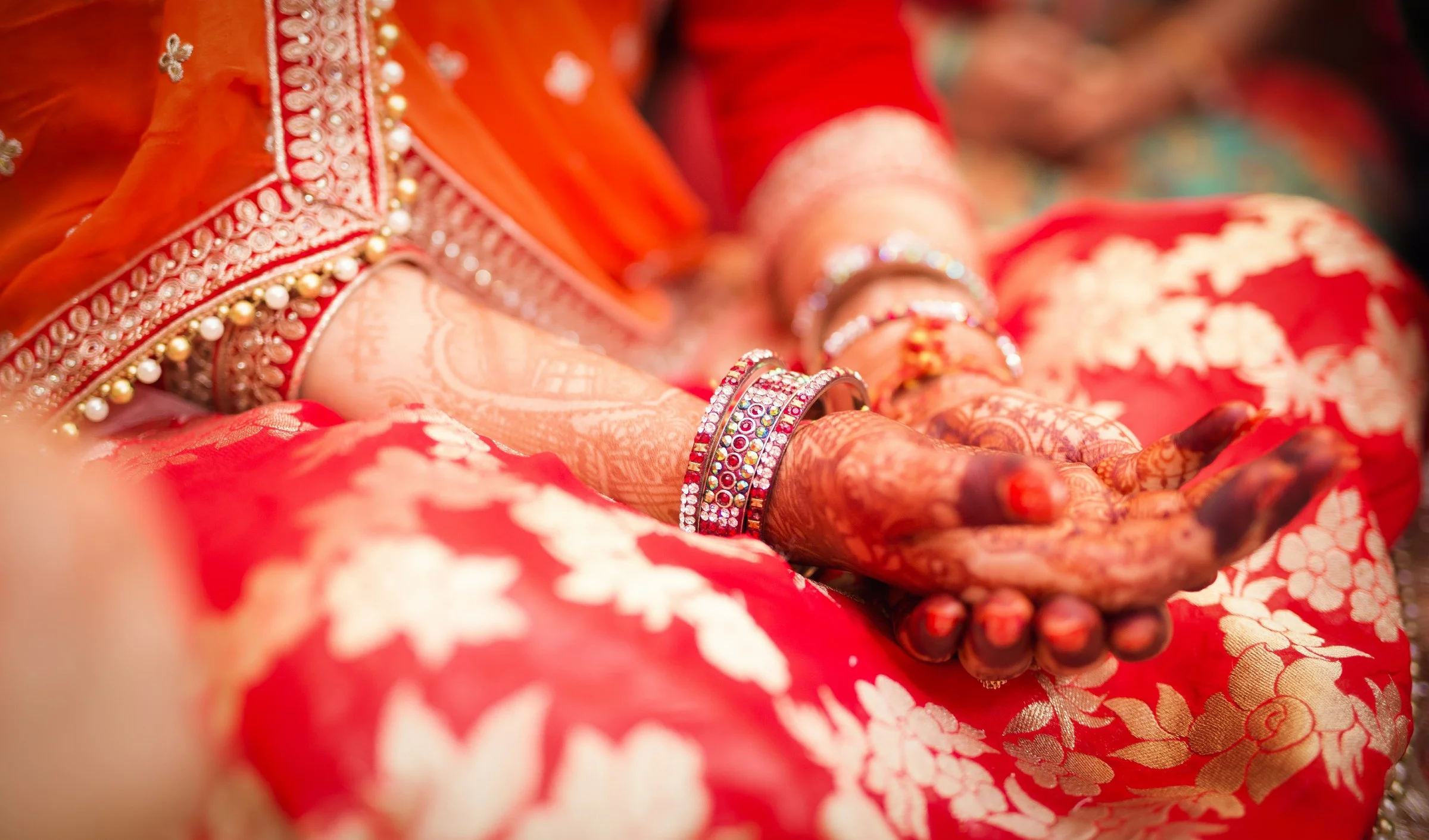 Close-up of a woman's hands adorned with colorful bangles and intricate henna designs, resting on a red and white floral fabric, wearing traditional Indian attire with detailed embroidery and jewelry.
