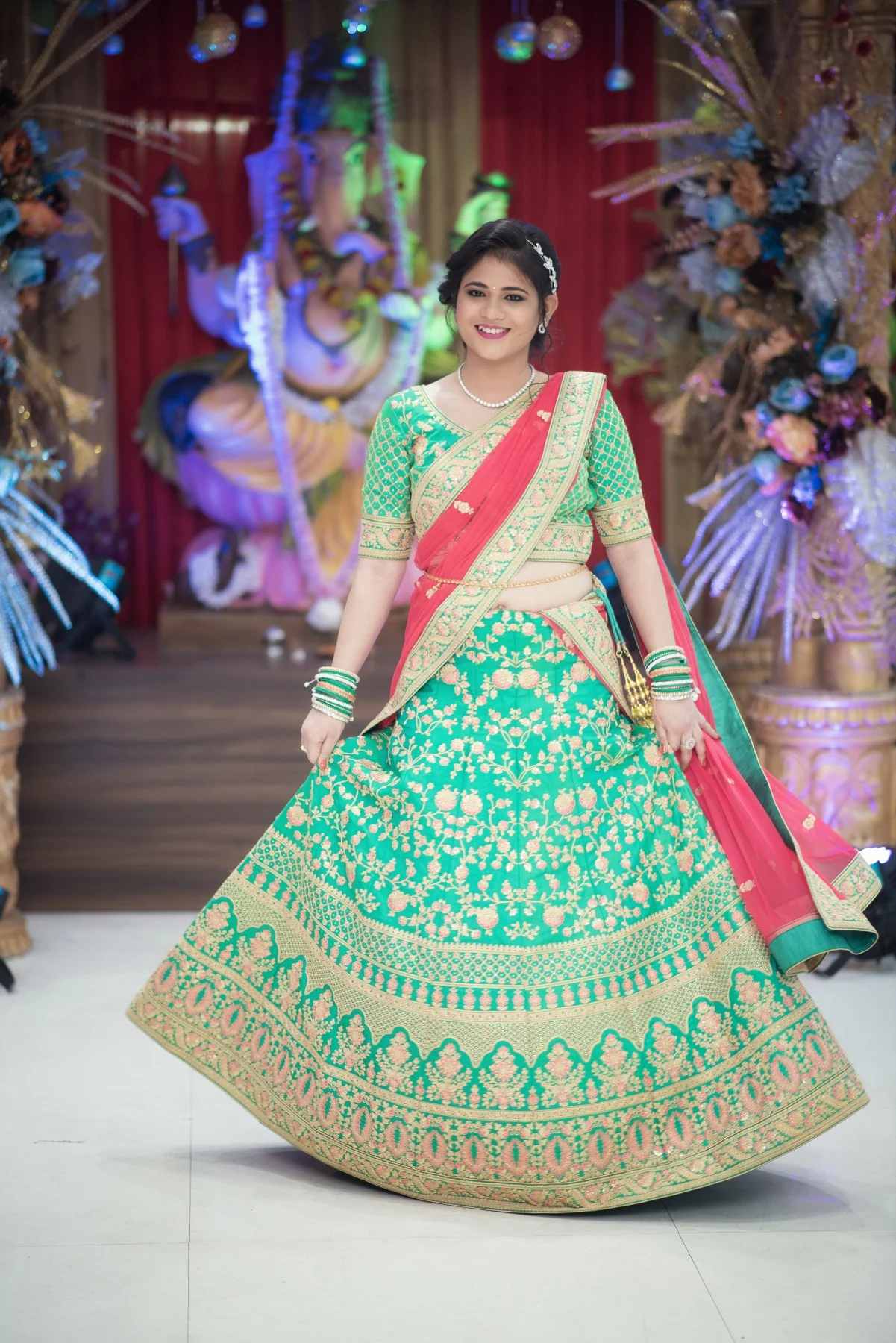 A woman dressed in traditional Indian attire, a green and gold lehenga with pink accents, standing in front of a colorful backdrop with a large Ganesh idol and floral decorations.