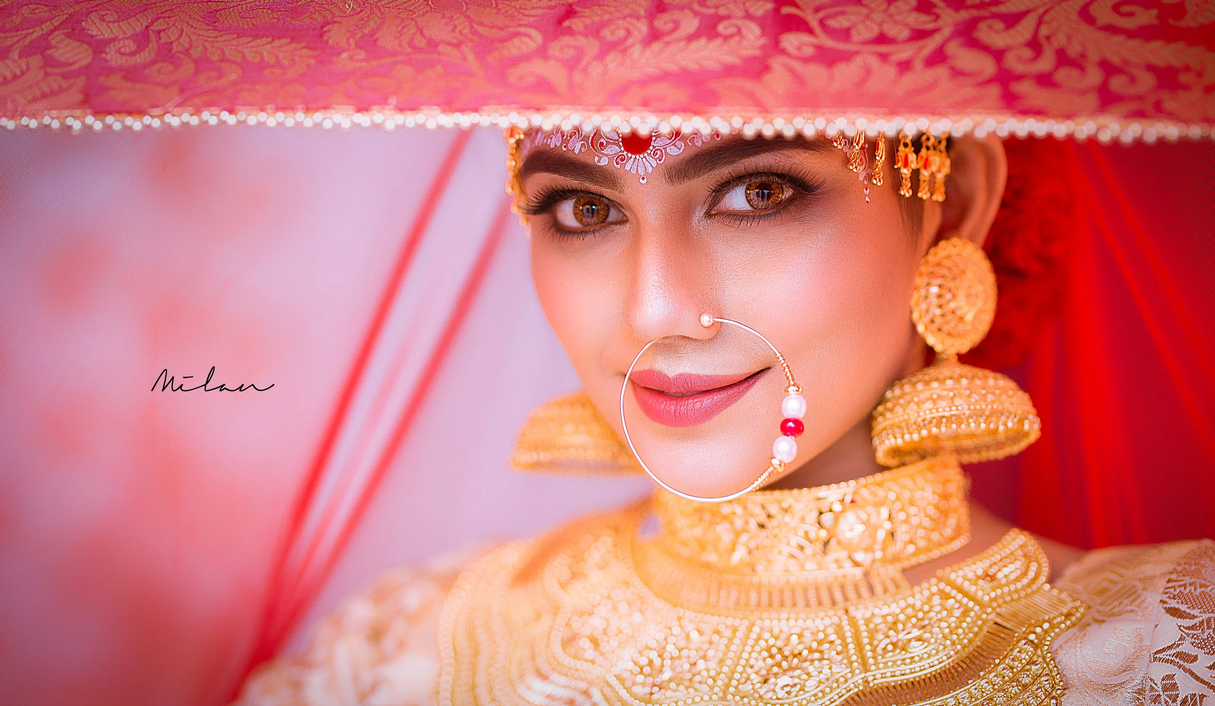Close-up of a woman wearing traditional Indian bridal jewelry and attire, with elaborate gold earrings, a nose ring, and intricate gold embroidery on her outfit, partially obscured by a decorative pink umbrella, looking directly at the camera with a 