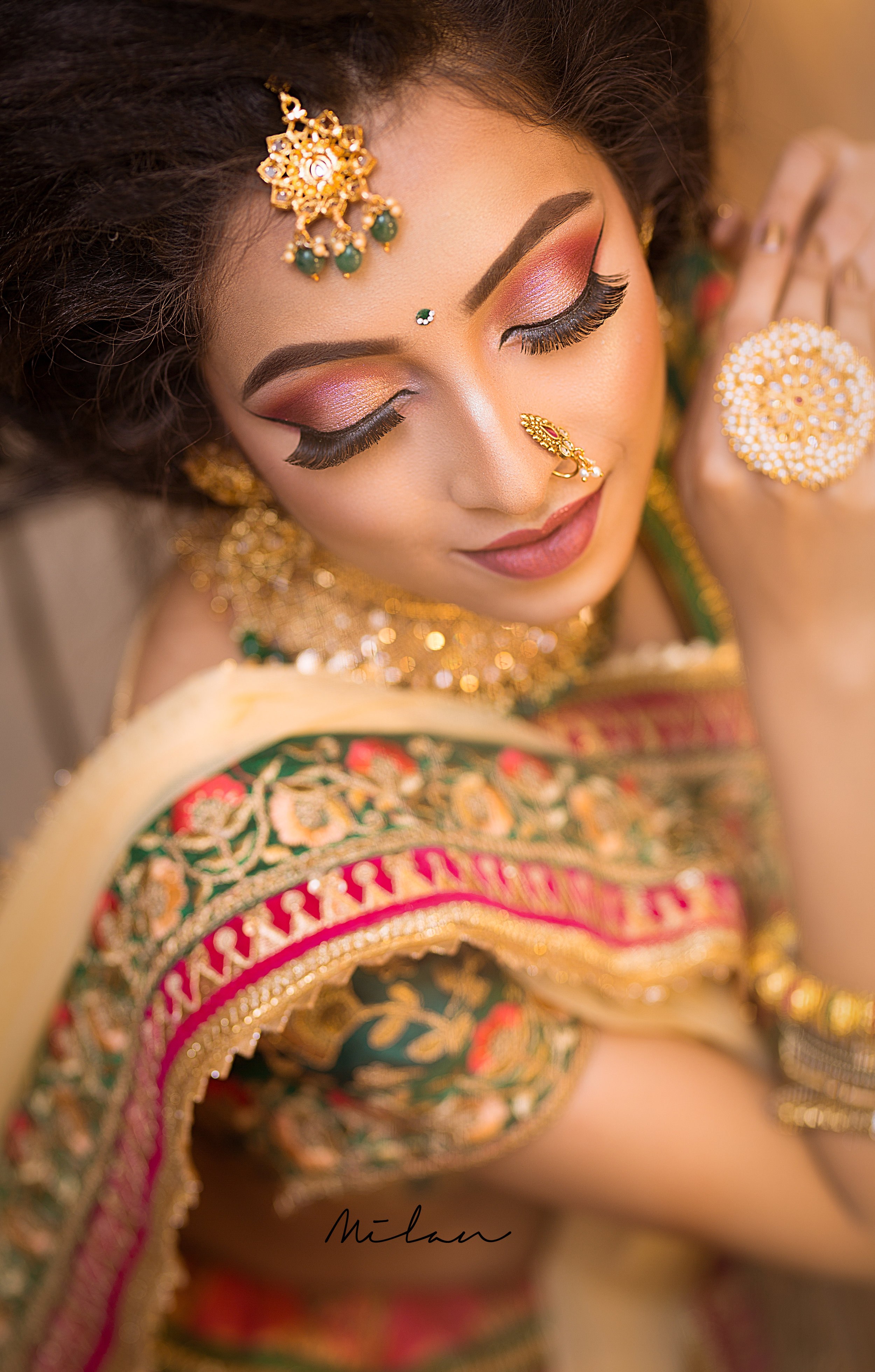 A woman dressed in traditional Indian attire with elaborate jewelry, including earrings, nose ring, and necklace, and vibrant makeup with pink and purple eyeshadow, long eyelashes, and nude lipstick, lying with her head tilted slightly downward and e