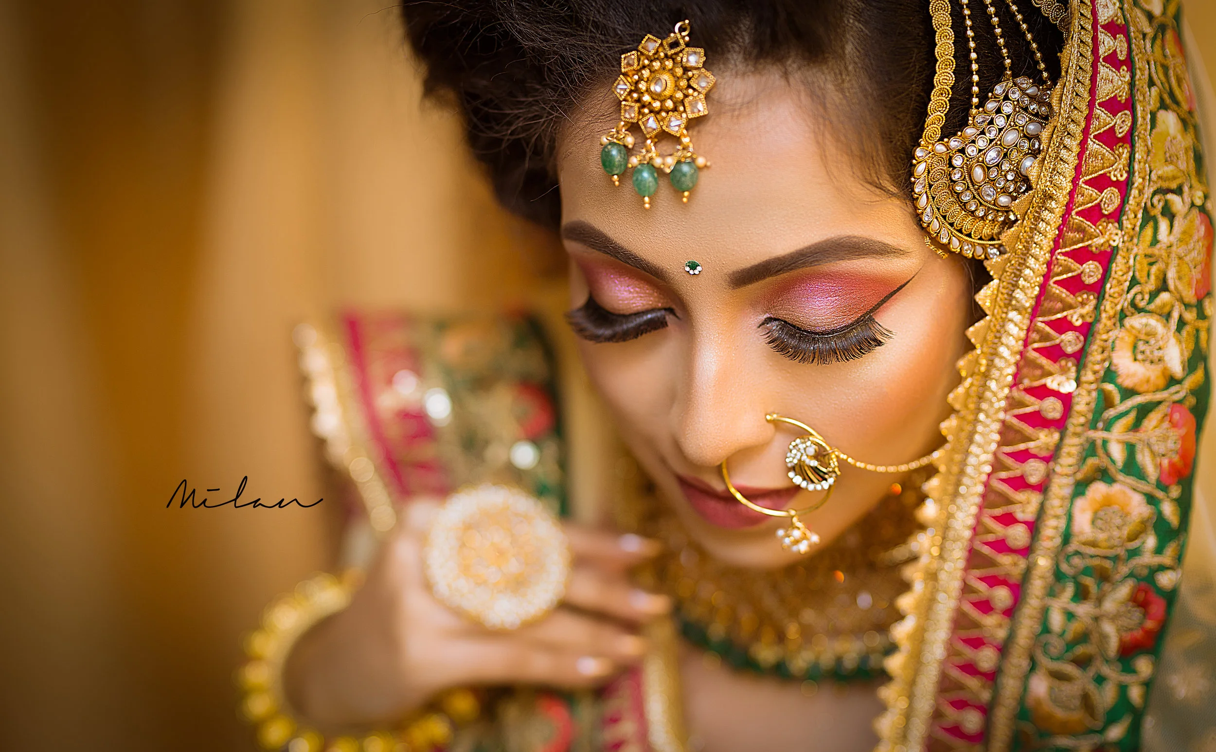 Close-up of a bride with traditional Indian jewelry and makeup, looking downward with eyes closed, wearing a richly embroidered red and gold saree