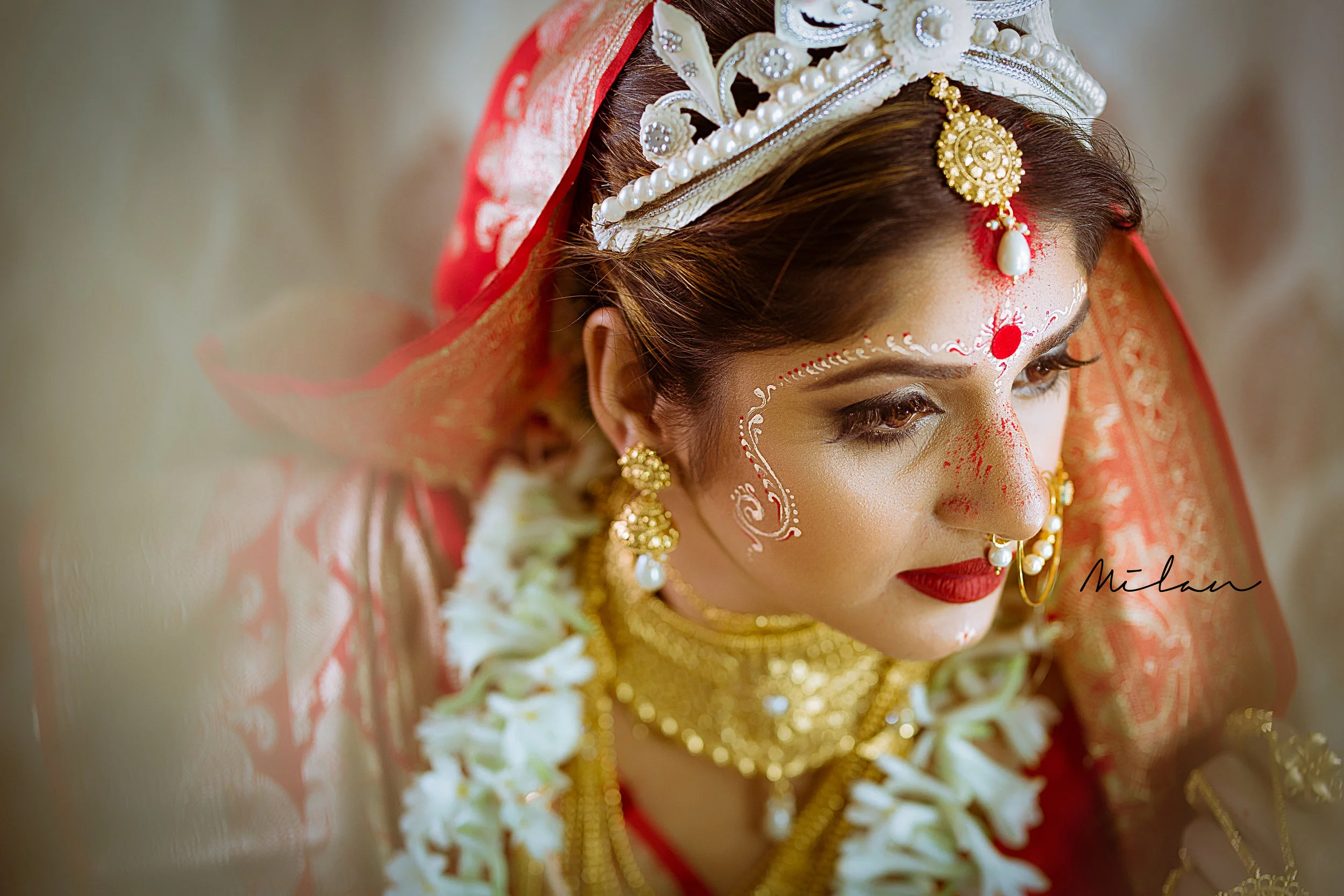 Close-up of a bride with traditional Indian wedding makeup and jewelry, including gold earrings, necklace, and headpiece, wearing a red and gold saree with floral garlands and white paint designs on her face.