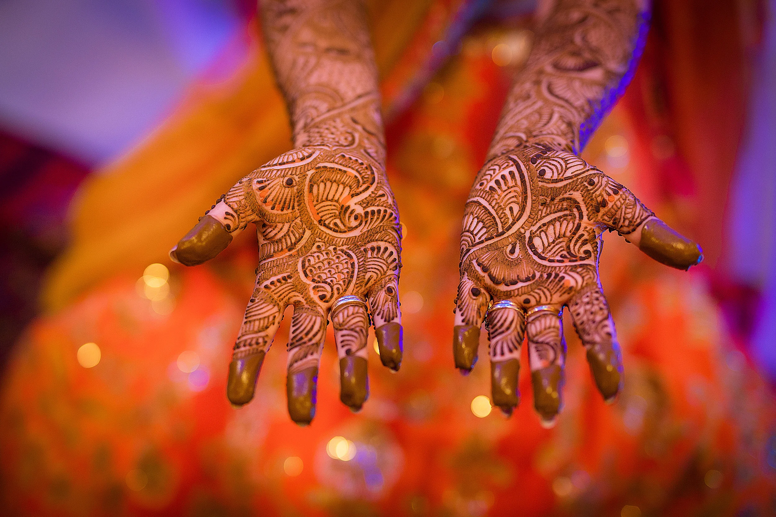 A close-up of hands decorated with intricate henna designs, with rings on some fingers, and painted nails, against a colorful blurred background.