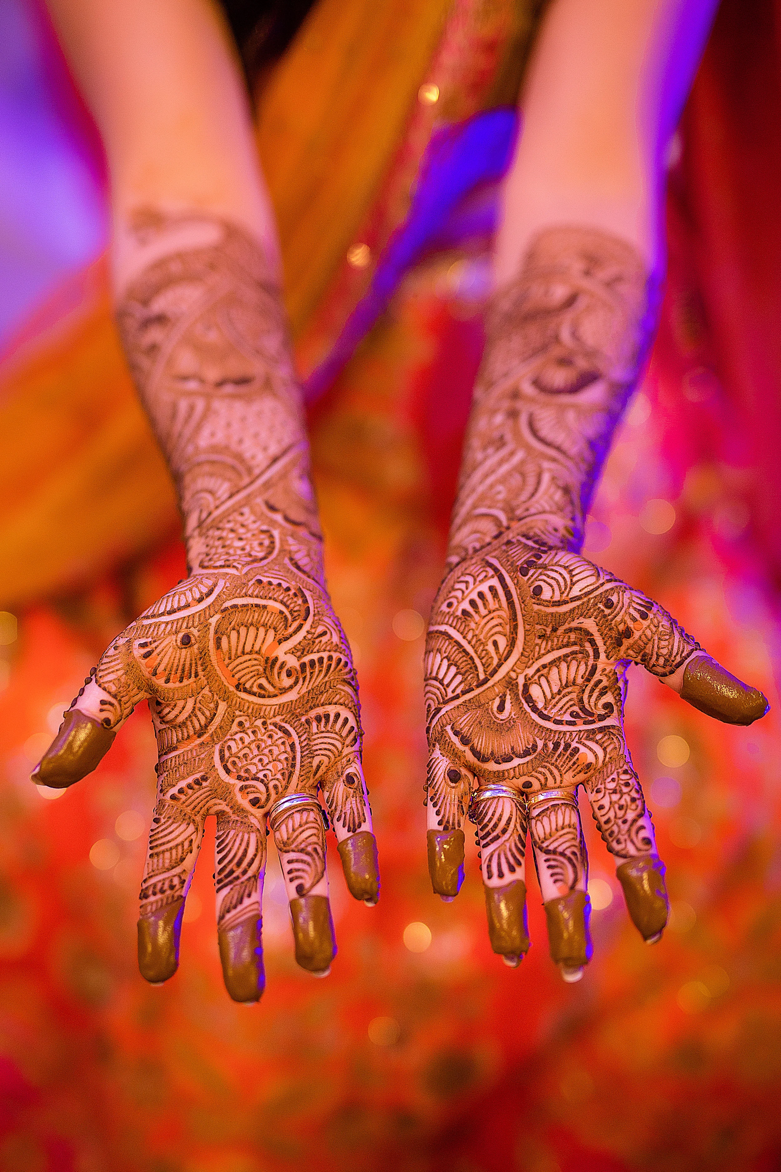 Close-up of hands with intricate traditional henna tattoo designs, adorned with rings, against a vibrant, colorful background.