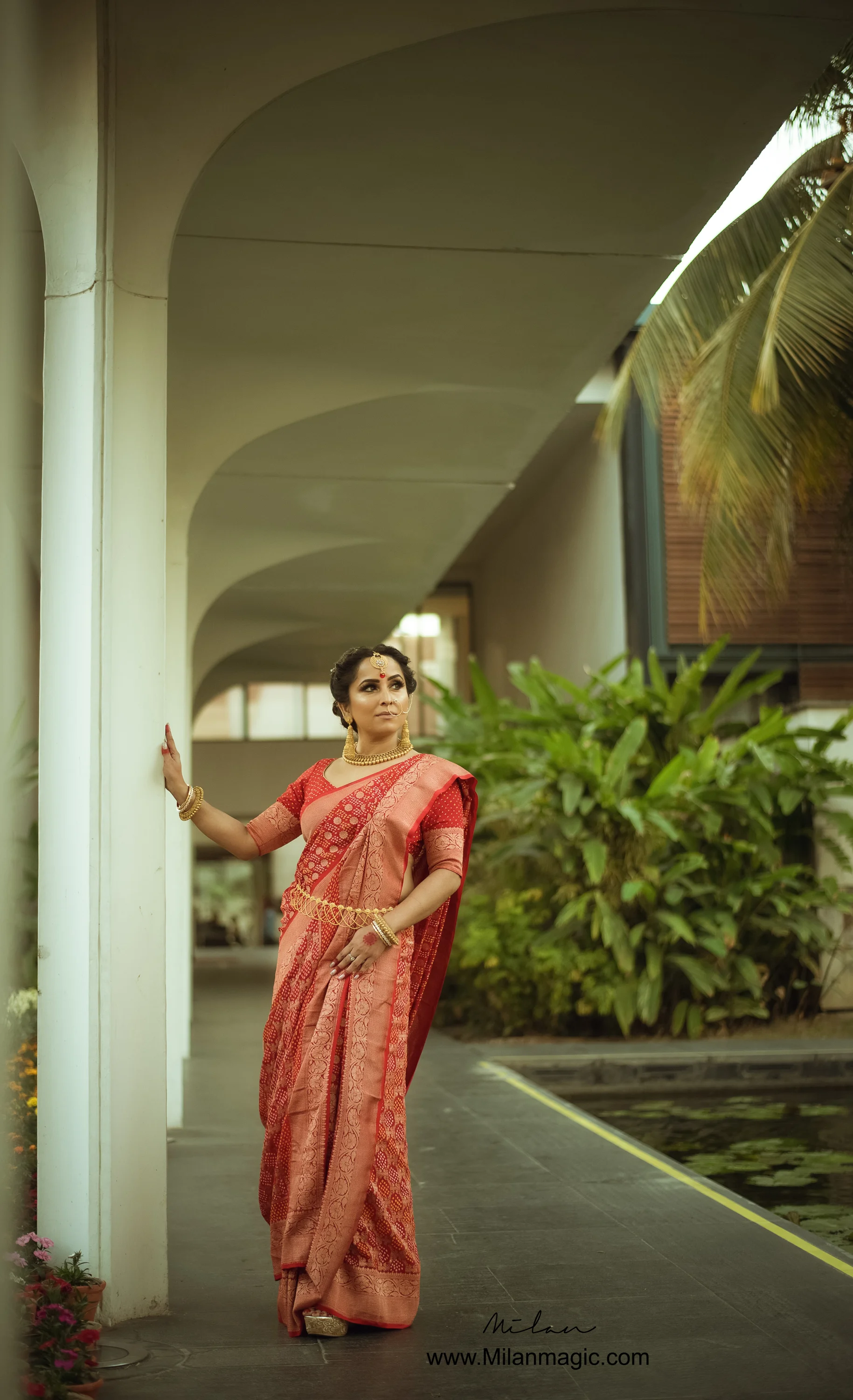 A woman dressed in a traditional red and gold saree standing on a walkway next to a white column, with lush green plants and a pond nearby.