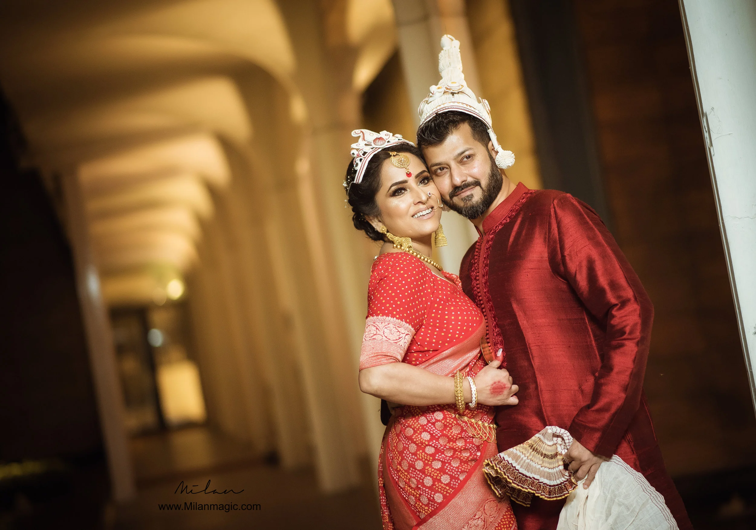 A couple dressed in traditional Indian wedding attire, with the woman in a red saree and ornate jewelry, and the man in a matching red kurta with a turban, standing closely together in a warmly lit corridor.