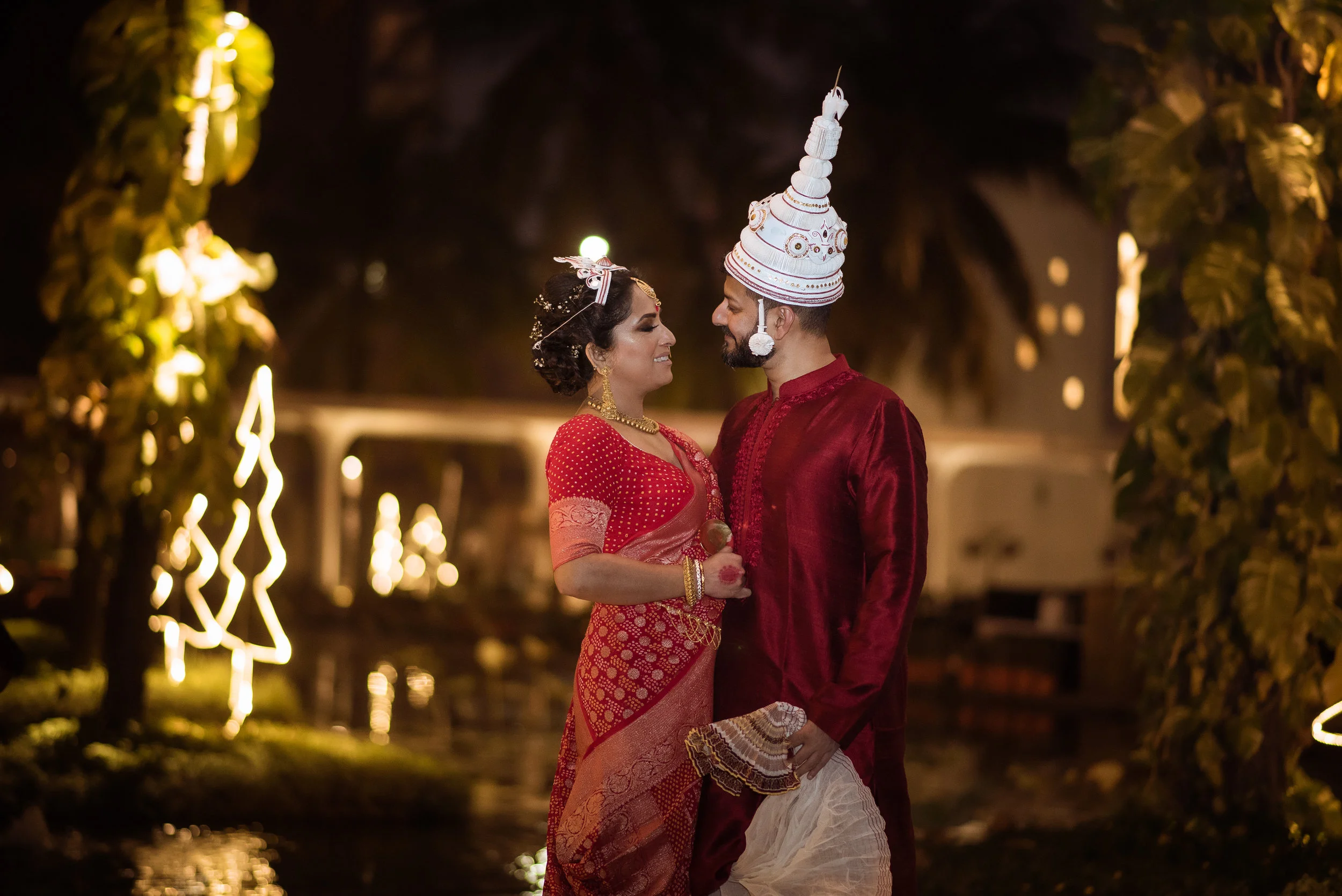 A couple dressed in traditional Indian wedding attire standing close and smiling at each other at night, surrounded by illuminated decorative lights.