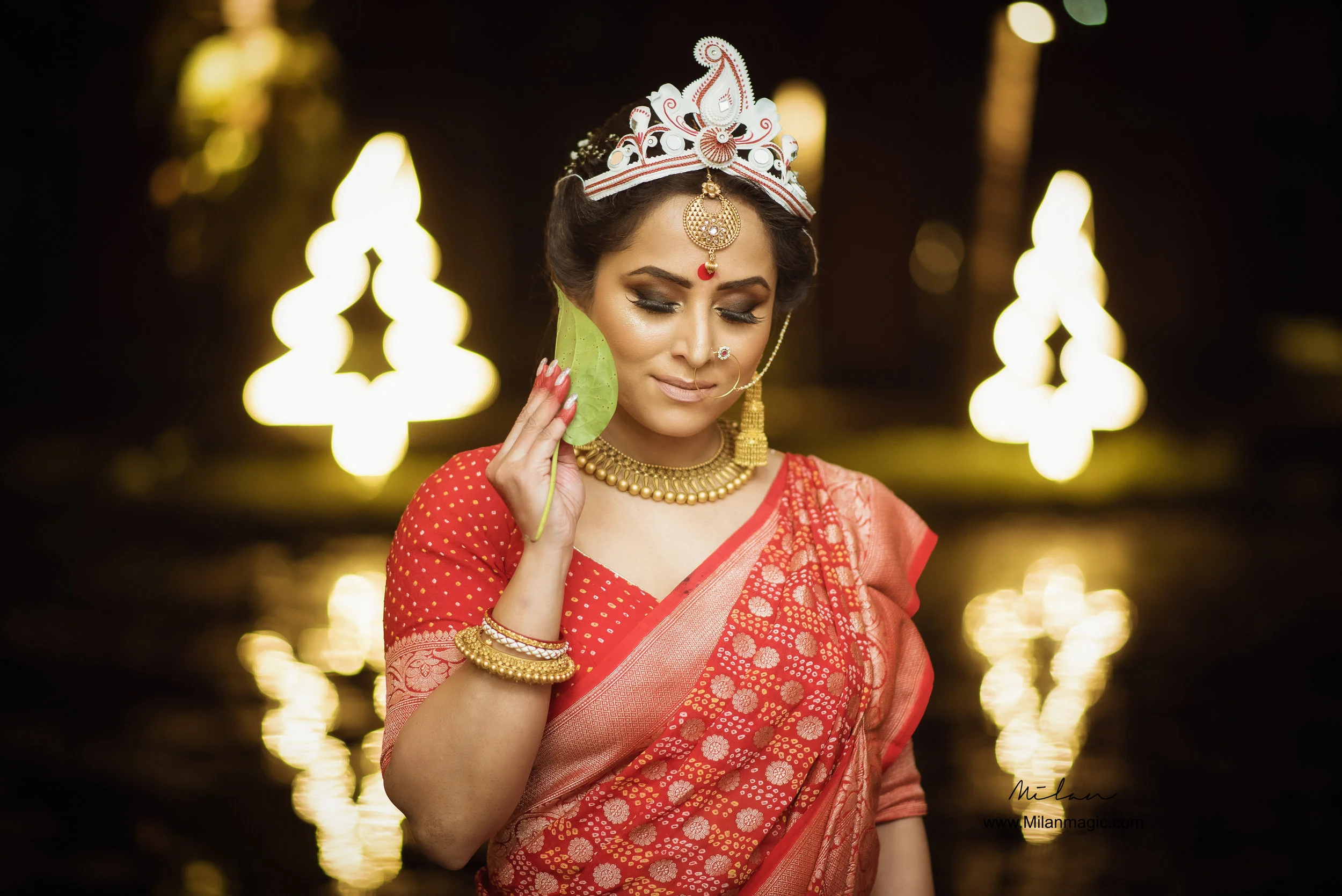 A woman dressed in traditional Indian attire holding a green leaf to her face, wearing jewelry and a crown, standing in front of illuminated decorative lights reflected in water.